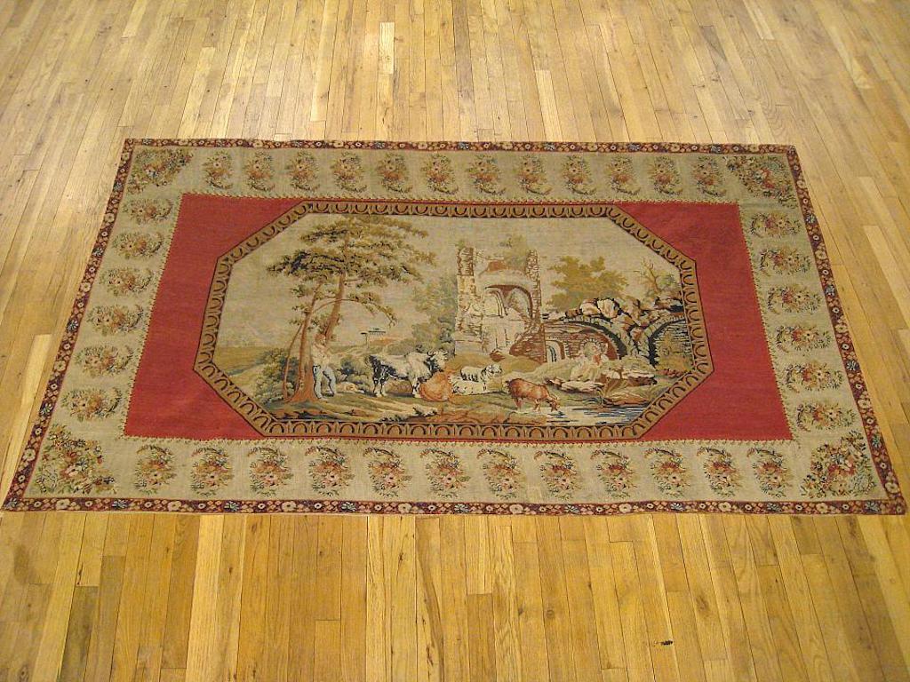 An English pictorial needlepoint tapestry from the 19th century, featuring a pastoral scene within a pendant window, with a farmer tending to his animals as they graze on a river bank before large arches. Enclosed within a main border of repeating