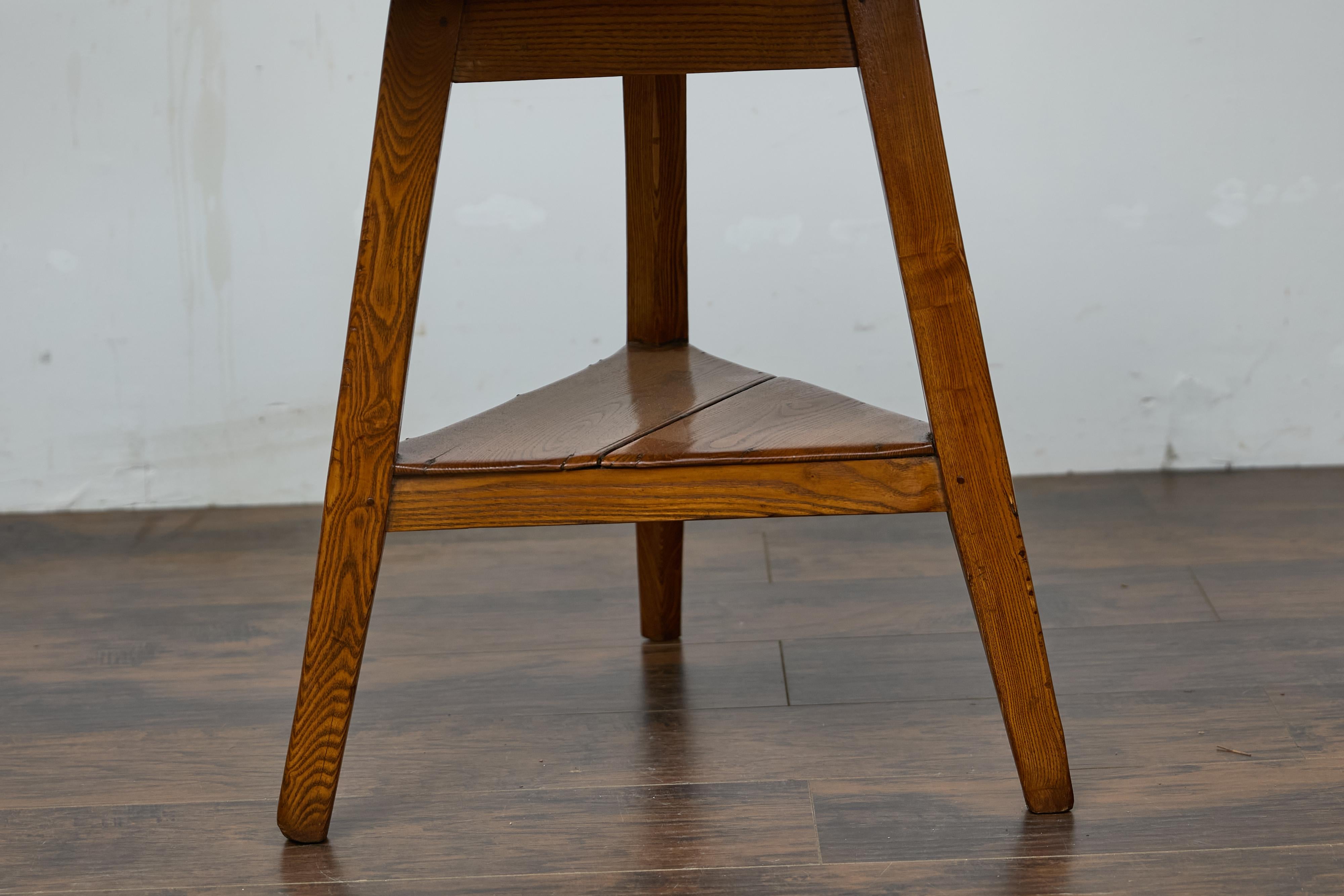 An English pine cricket table from the 19th century with circular top, tripod base and triangular shelf. This English pine cricket table from the 19th century exudes rustic charm with its circular top and sturdy tripod base. The top, composed of