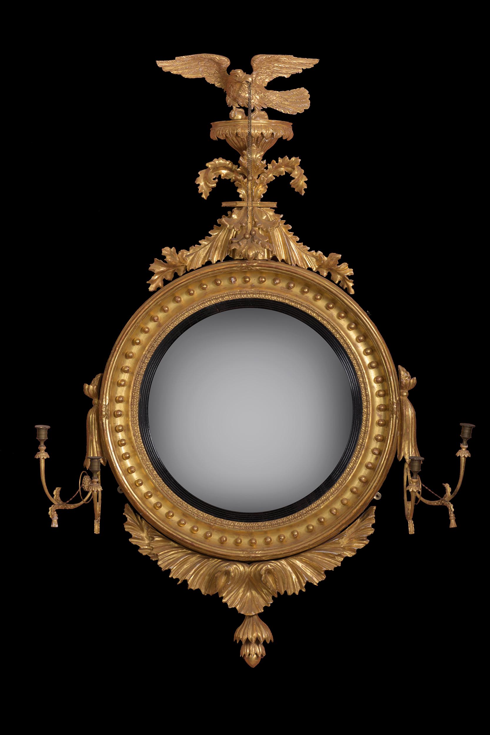 A very fine Regency period carved giltwood and Gesso convex mirror, the plate within a reeded ebonized slip with ball decoration surmounted by an Eagle pediment. It has sconces to either side with the original gilt brass candleholders and sunburst
