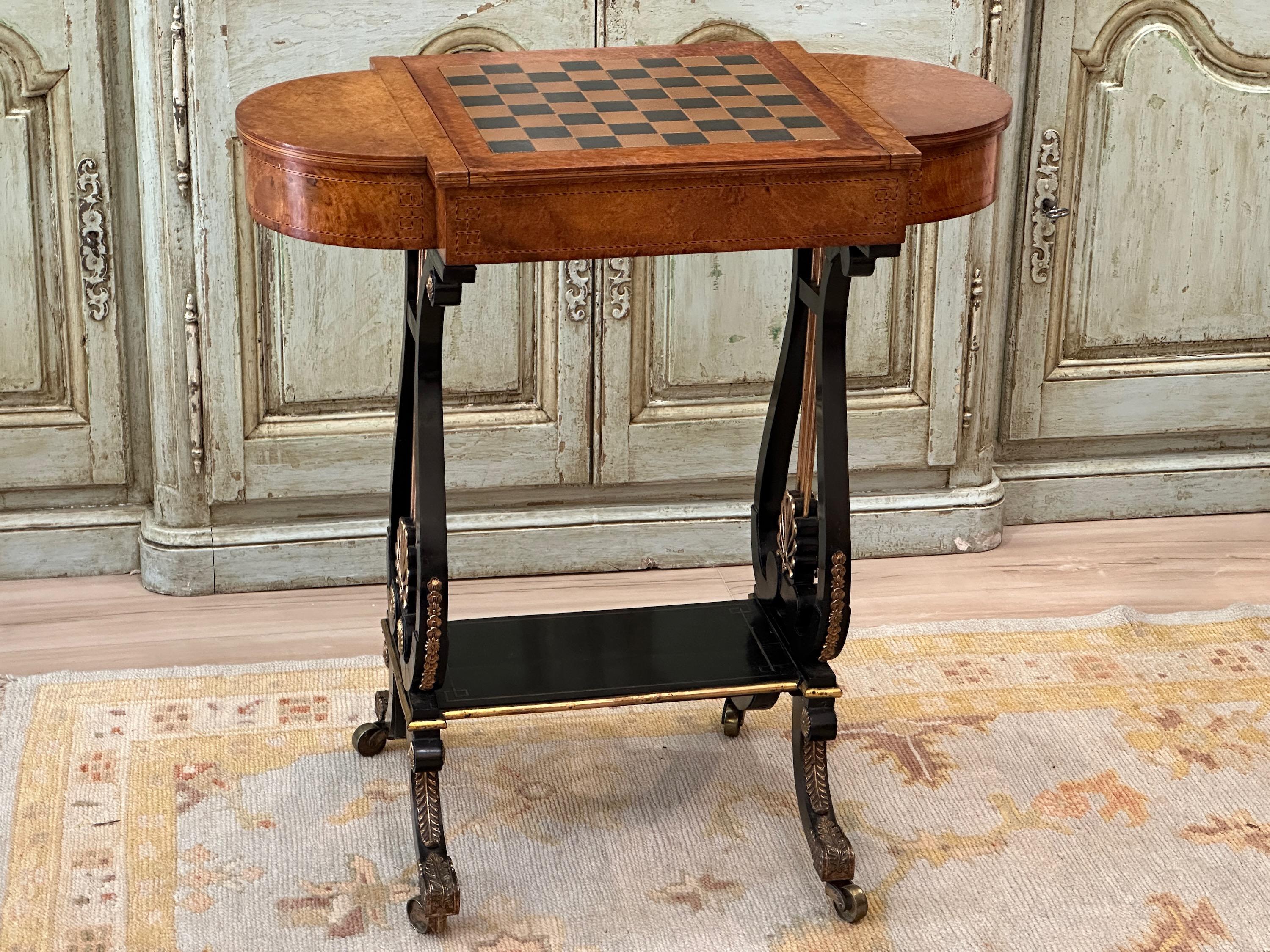 An early 19th C. English Regency period games table in amboyna burl wood with inset tooled leather backgammon board and tooled chess board on reverse side of top resting on ebonized lyre shaped legs heavily adorned with gilt bronze acanthus shaped