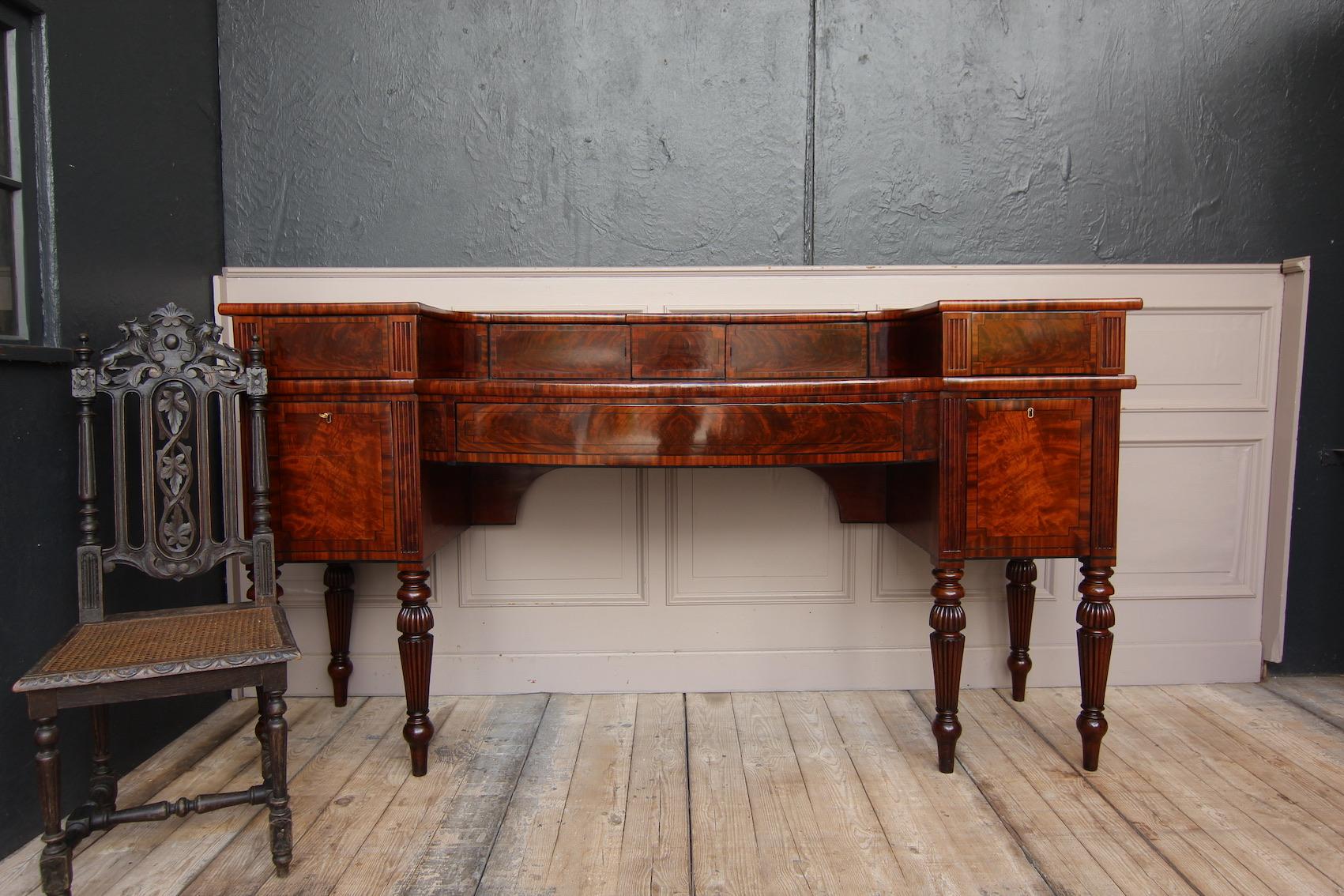 Credenza inglese Regency degli inizi del XIX secolo. Mogano massiccio e impiallacciato.

Il corpo a due livelli poggia su 6 gambe tornite, la cui parte anteriore è prevalentemente impiallacciata in legno di mogano fiammato e decorata con intarsi