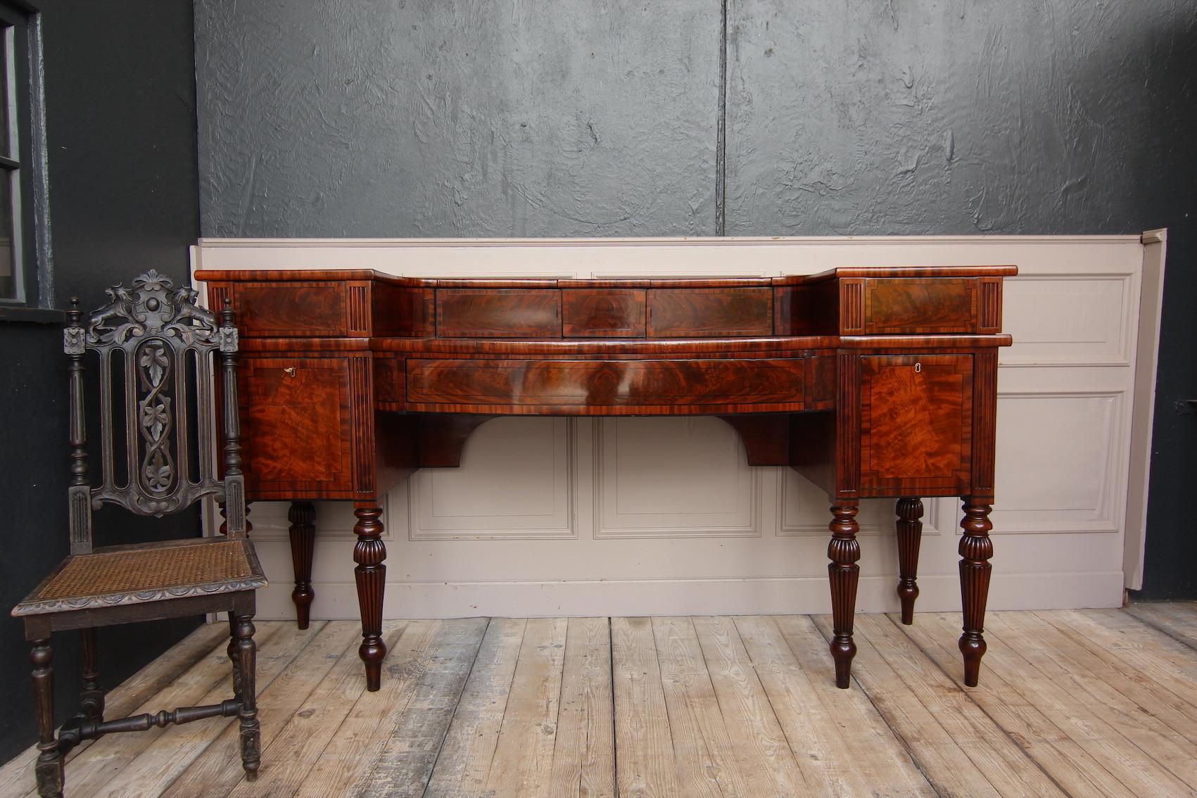 English Regency sideboard from the early 19th century. Solid mahogany and veneered.

Two-tier body standing on 6 turned legs, the front of which is predominantly veneered with flamed mahogany wood and decorated with ebony thread inlays.
A large,