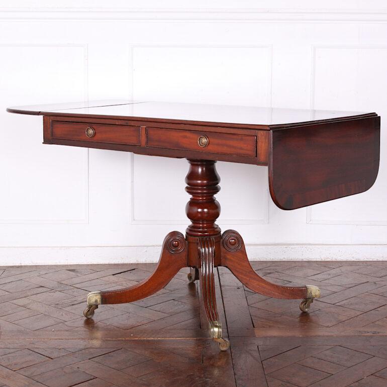 English mahogany Regency sofa table in figured mahogany, the top and drop ends with mahogany banded edges, and fitted with a pair of drawers to the apron and ‘dummy’ drawers to the reverse. Standing on a turned mahogany pedestal support above four