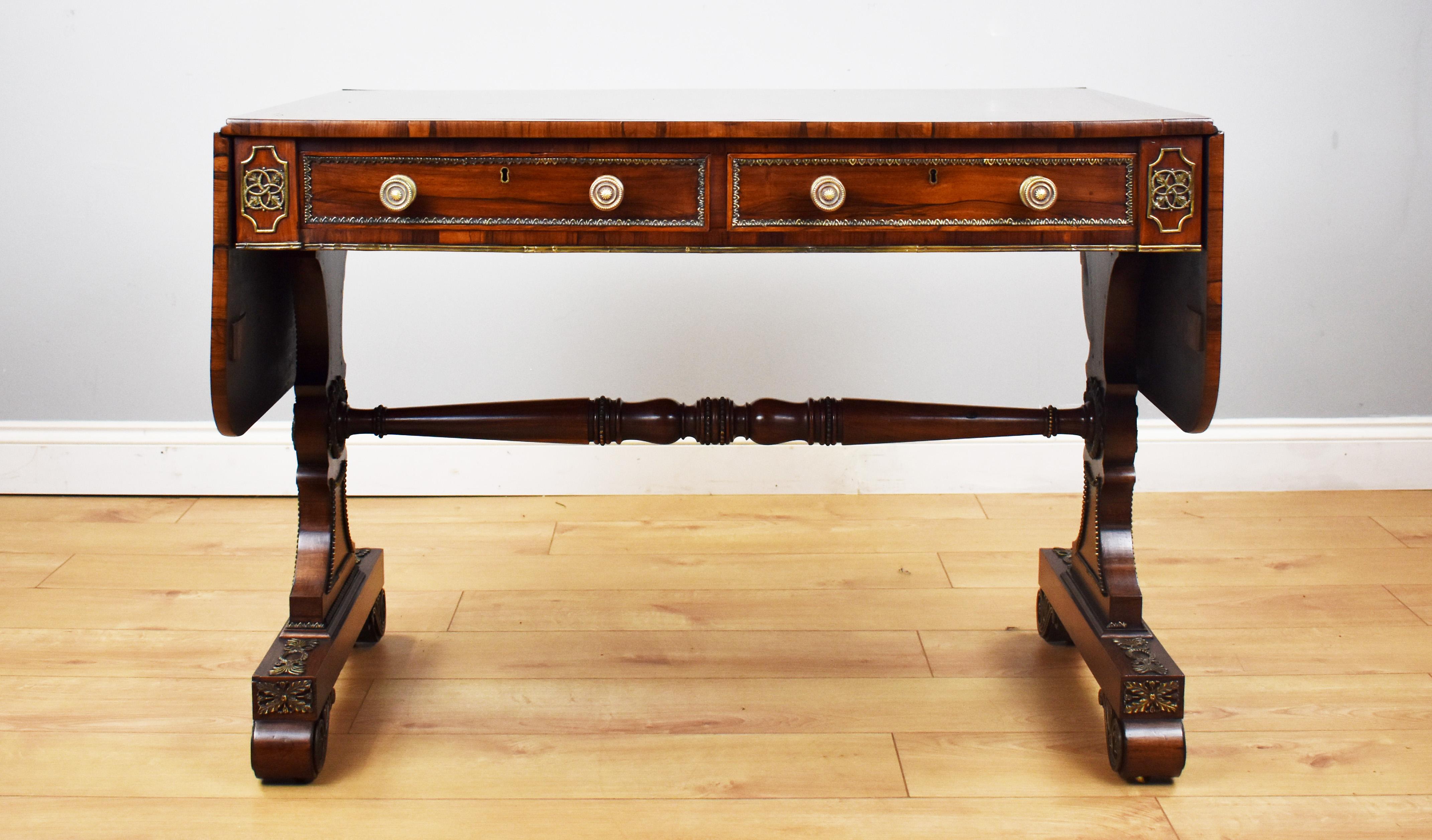 For sale is a fine quality Regency period rosewood and brass inlaid sofa table, having drop-leaf ends outlined with brass fleur-de-lis inlay over a pair of frieze drawers with brass mouldings and brass handles, with two dummy drawers on the opposing