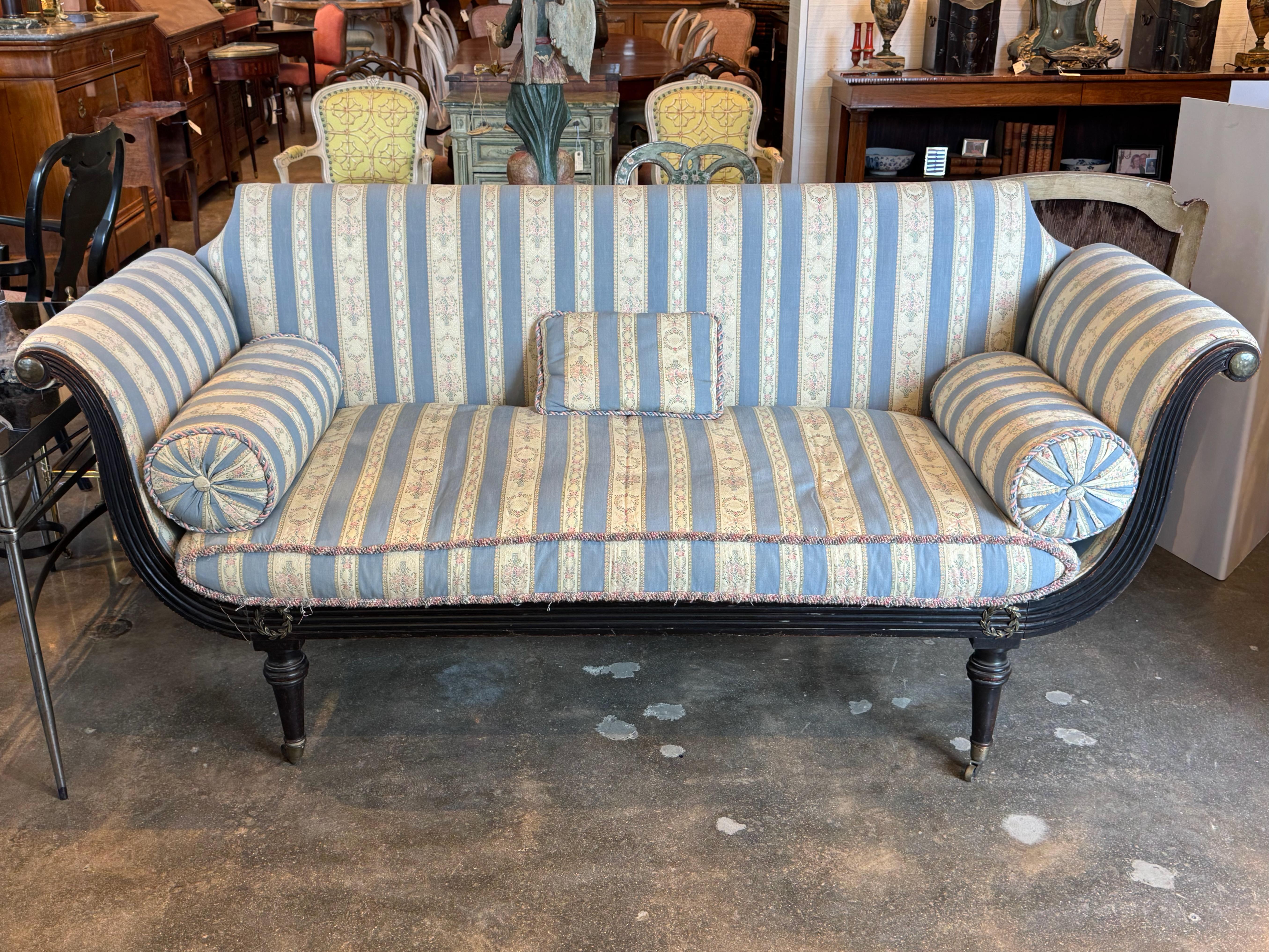 19th century English Regency-style sofa with a gracefully curved frame and scrolled arms, finished in dark wood. Upholstered in striped fabric with soft blue, cream, and floral bands, accented by rosettes and corded trim. Includes two bolster
