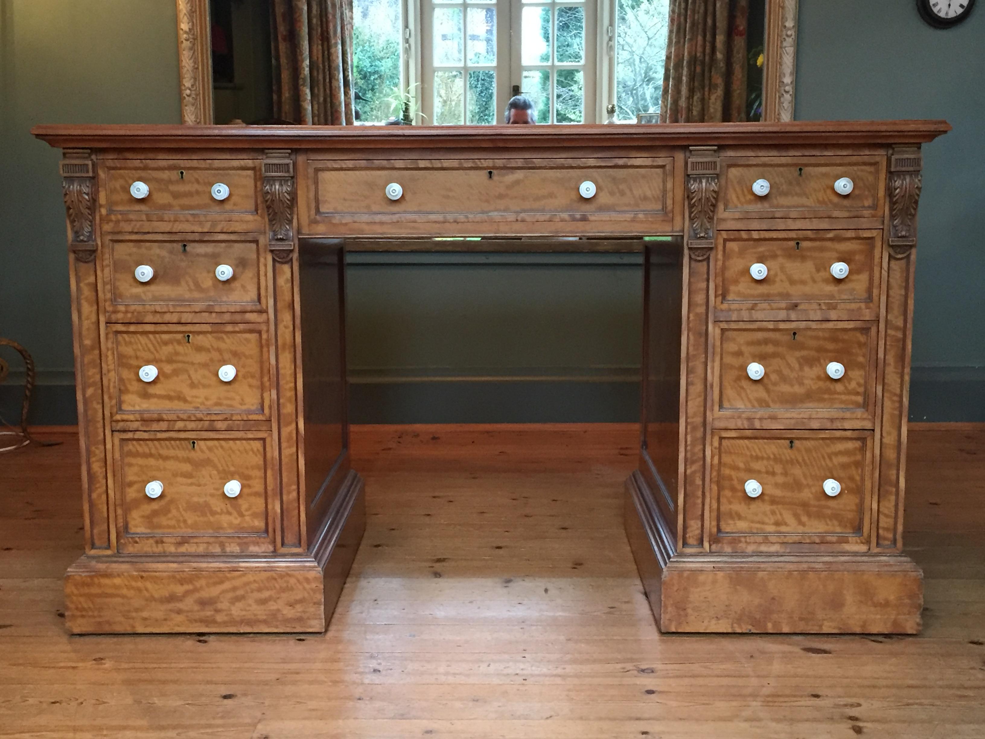 An exquisite 19th century pedestal desk in satinwood, the original tooled leather top over a central frieze drawer; each pedestal with carved corbels, four graduated drawers all with their original handles and plinth bases. This desk is in one