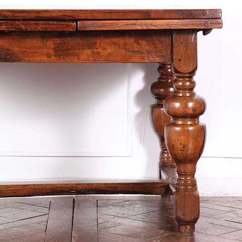 English rustic solid oak plank-top draw-leaf table standing on boldly-turned legs united by an H-stretch at the base. Width dimension shown is with leaves drawn out. Table is 48