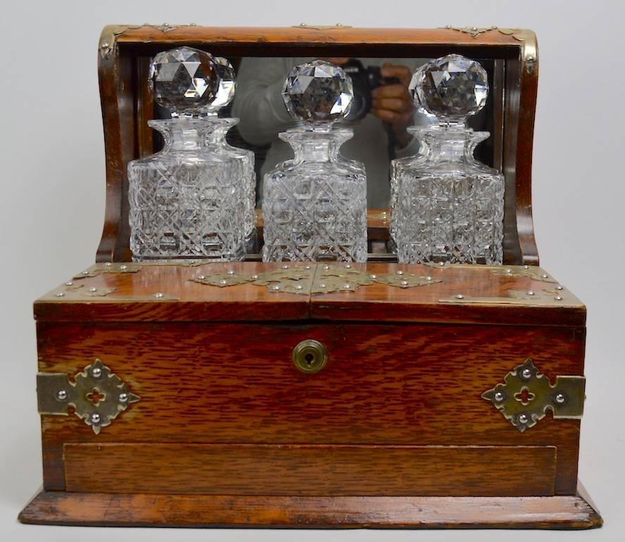 Nice oak case tantalus decanter set by Branah London, with three crystal decanters. The case shows some minor cosmetic wear, is missing one decorative brass element, and we do not have the key to open the lower storage drawer, all bottles are in