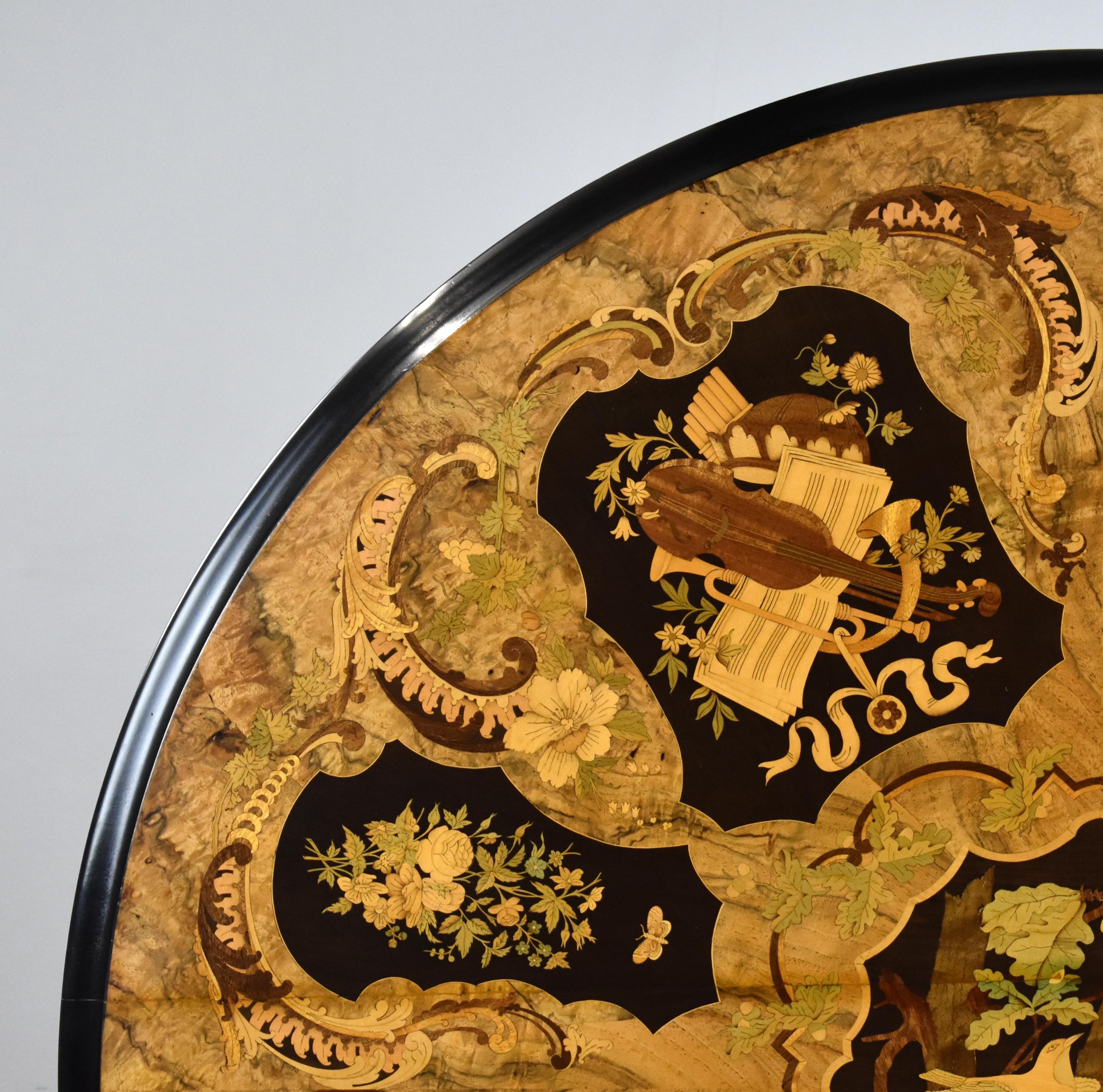 Il s'agit d'une table de petit déjeuner circulaire en ronce de noyer et marqueterie de style victorien anglais. Le plateau est abondamment incrusté de marqueterie florale et de panneaux représentant divers instruments de musique, au-dessus d'une