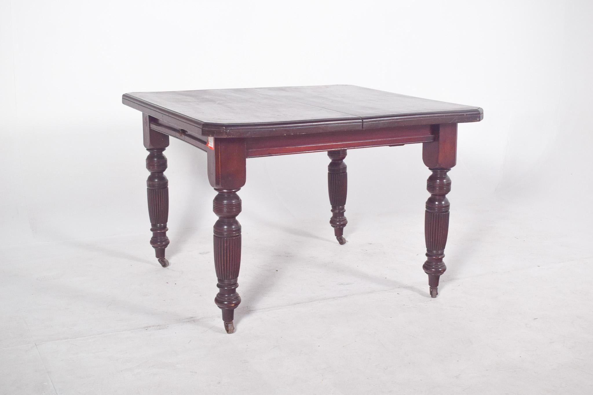 English dining table. It has a square top with slightly jagged edges, a smooth surface and a darkened finish. It rests on four turned baluster legs decorated with vertical fluting with small metal wheels, making it easy to move. The piece has a