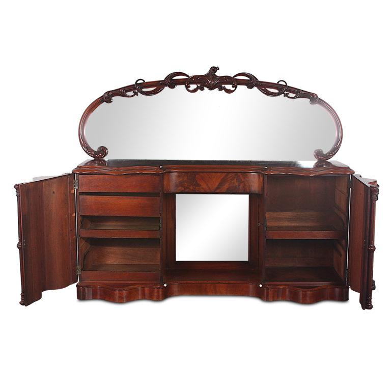 A dramatically-shaped and carved English Victorian sideboard with curved flame mahogany veneer paneled doors to each pedestal and a highly-carved mirrored back. Pedestals open to reveal fitted shelves and drawers and there is a single drawer fitted