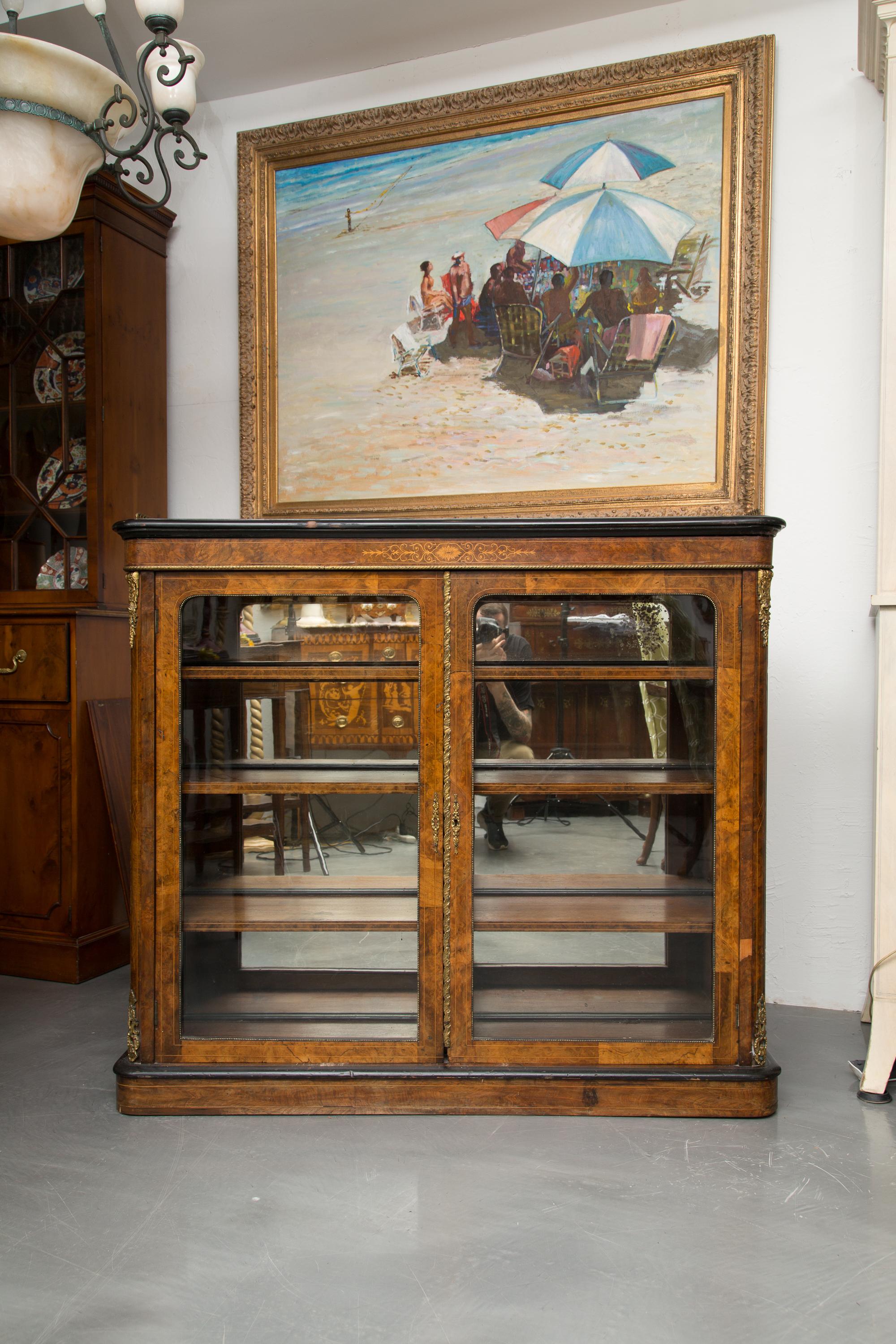 This is as very simple English Victorian open walnut bookcase. The top has an ebonized boarder over an inlaid frieze and two glass-inset cabinet doors revealing three interior shelves fronting a mirrored back. The case piece is situated on a plinth