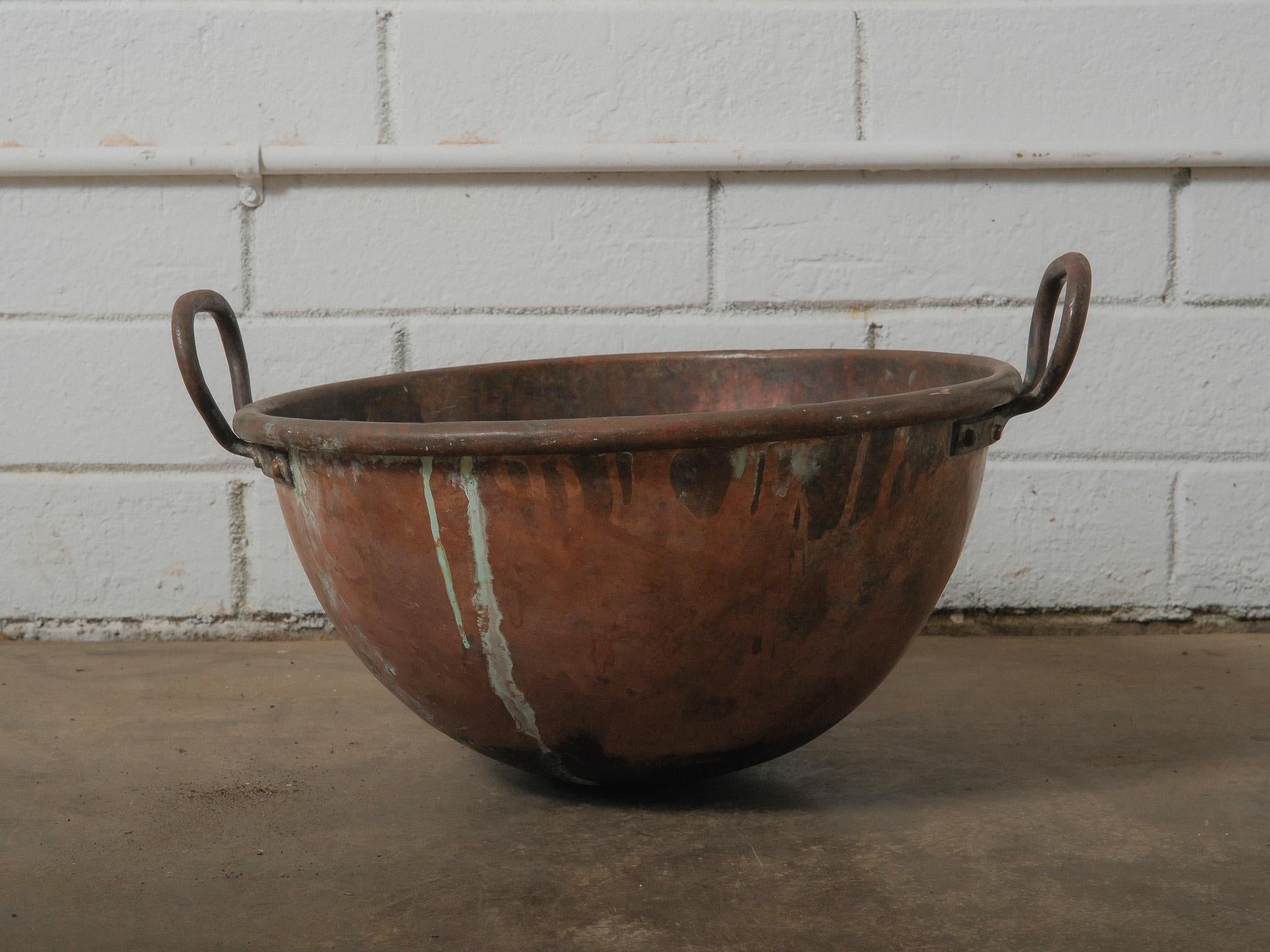 A substantial hand-formed copper vat with a deep rounded profile and expressive surface character. The vessel shows a warm, time-softened copper tone layered with areas of verdigris, oxidation, and dark burnishing from long and practical use. The