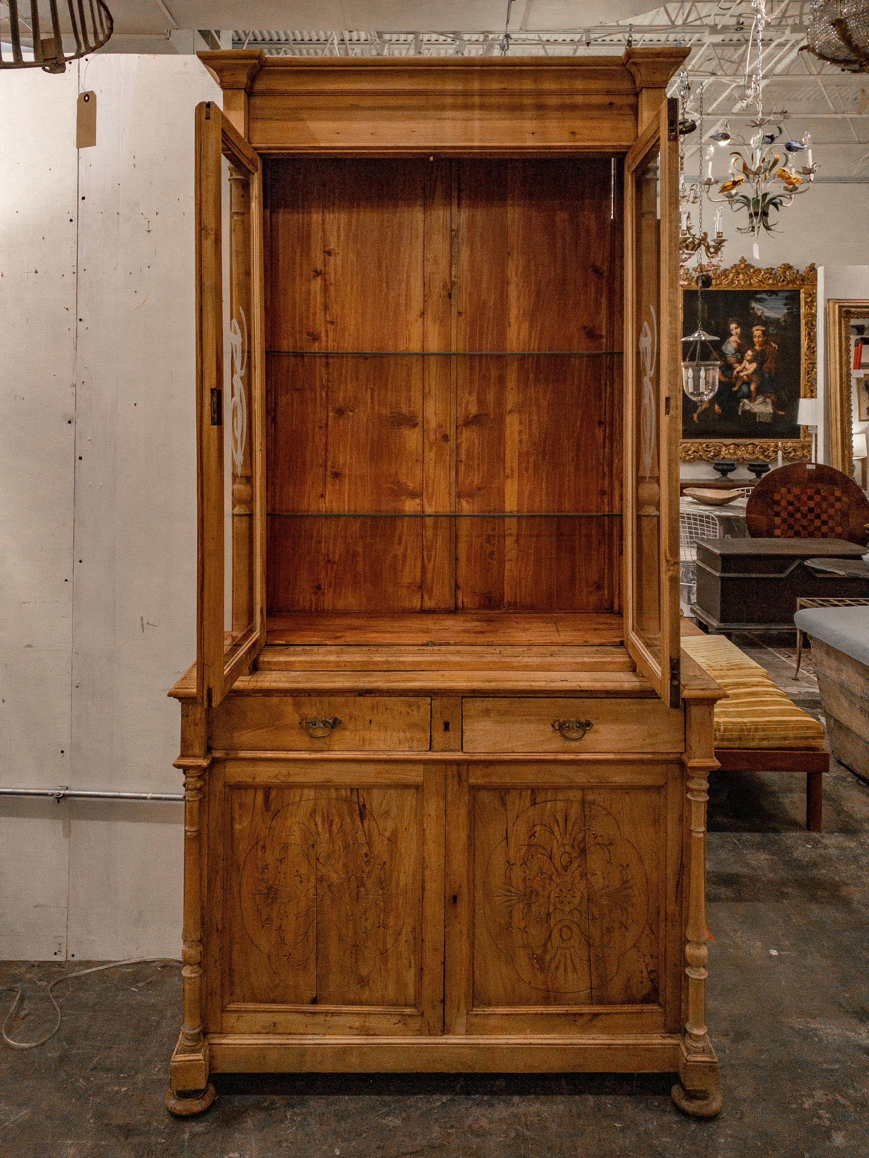 The 19th Century European Pine Bookcase exudes elegance and functionality through its exquisite craftsmanship and thoughtful design. Intricately carved details adorn its exterior, showcasing the skilled handiwork of the era. The two etched glazed