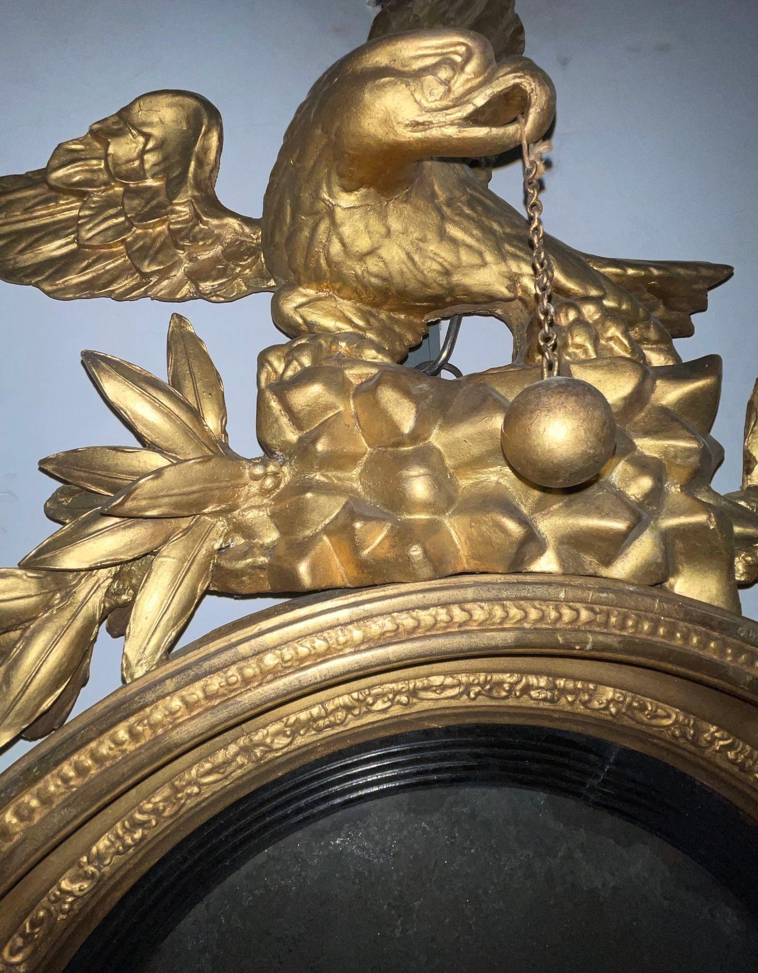 A magnificent American 19th-century Federal-style bullseye mirror, featuring a richly carved giltwood frame and a dramatic full-body eagle crest. The eagle stands majestically atop a rock and garland, holding a ball and chain in its beak,