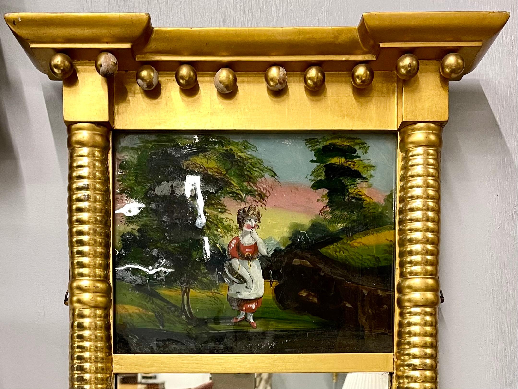 Un miroir mural ou de table finement détaillé, décoré d'un panneau d'églomisé en verre représentant une jeune fille dans une zone boisée, flanqué d'un cadre doré fin rappelant l'époque du fabricant. Can peut facilement être placé sur un support et