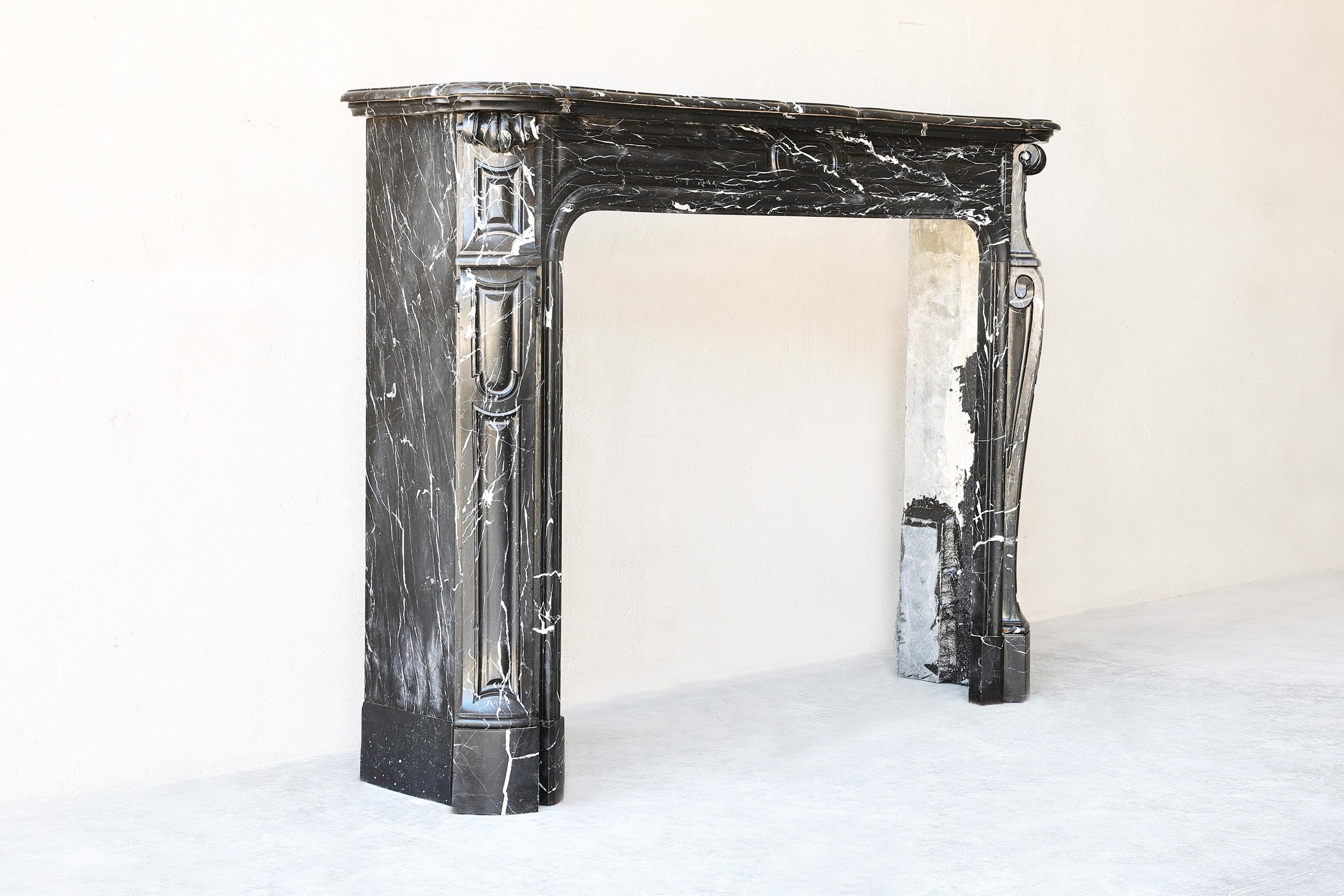 This 19th Century antique Pompadour fireplace, sculpted from Black Marquina marble, is a breathtaking masterpiece. This elegant relic boasts exquisite veining in shades of white and gray, which gracefully traverse the deep black backdrop. Its