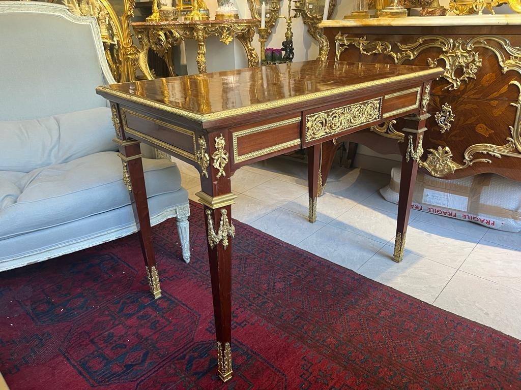 This Louis XVI-style writing desk from the Napoleon III period exudes exquisite craftsmanship thanks to its use of mahogany and opulent gilded bronze embellishments. Its tabletop is elegantly framed by a gilded bronze moulding featuring heart-shaped