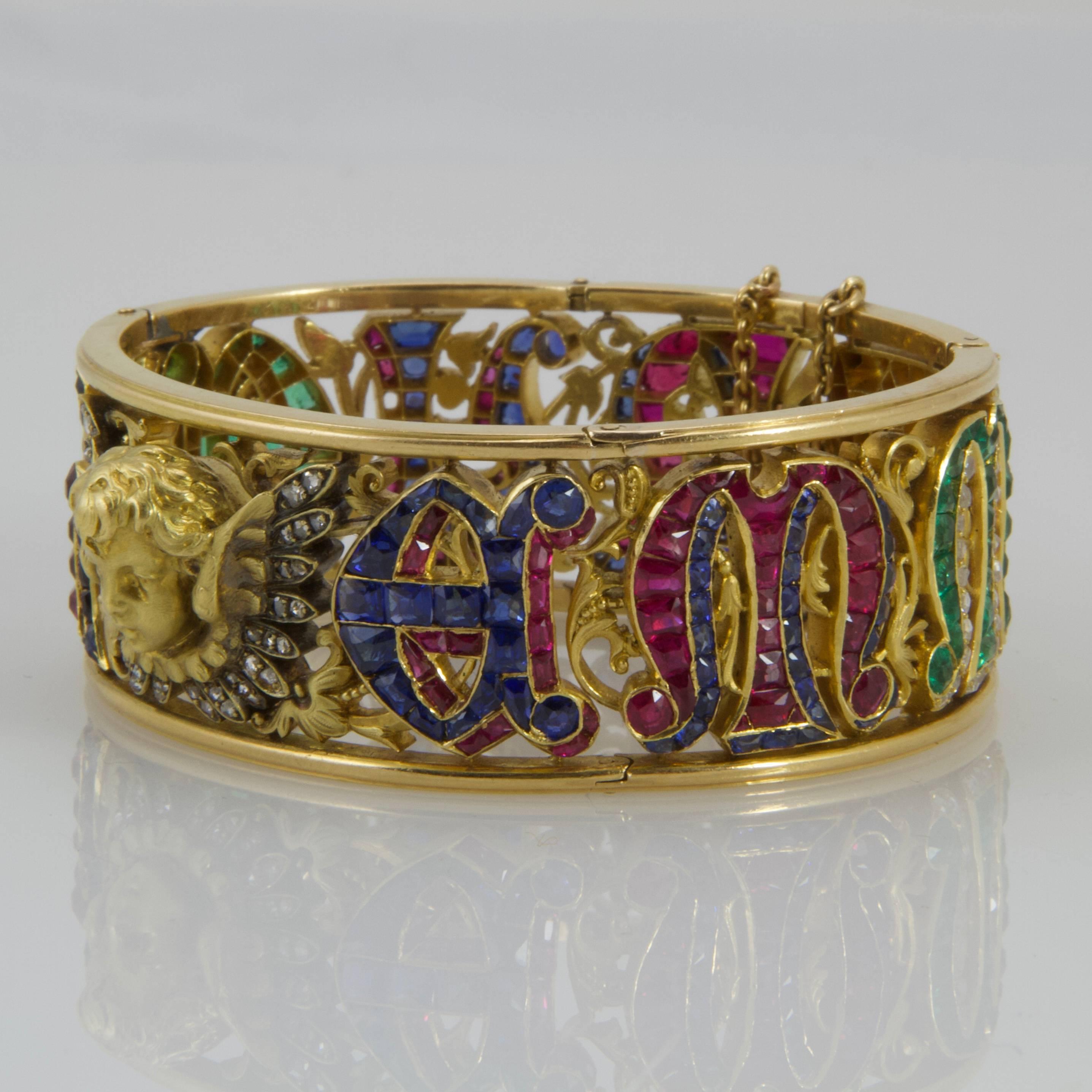 18kt yellow mat gold chiseled foliage pattern bangle presenting on the center a face of angel with rose cut diamond wings. Overall the bangle, the letter of Emmanuel are displayed in calibre precious stones, rubies shaded by sapphires, and vice