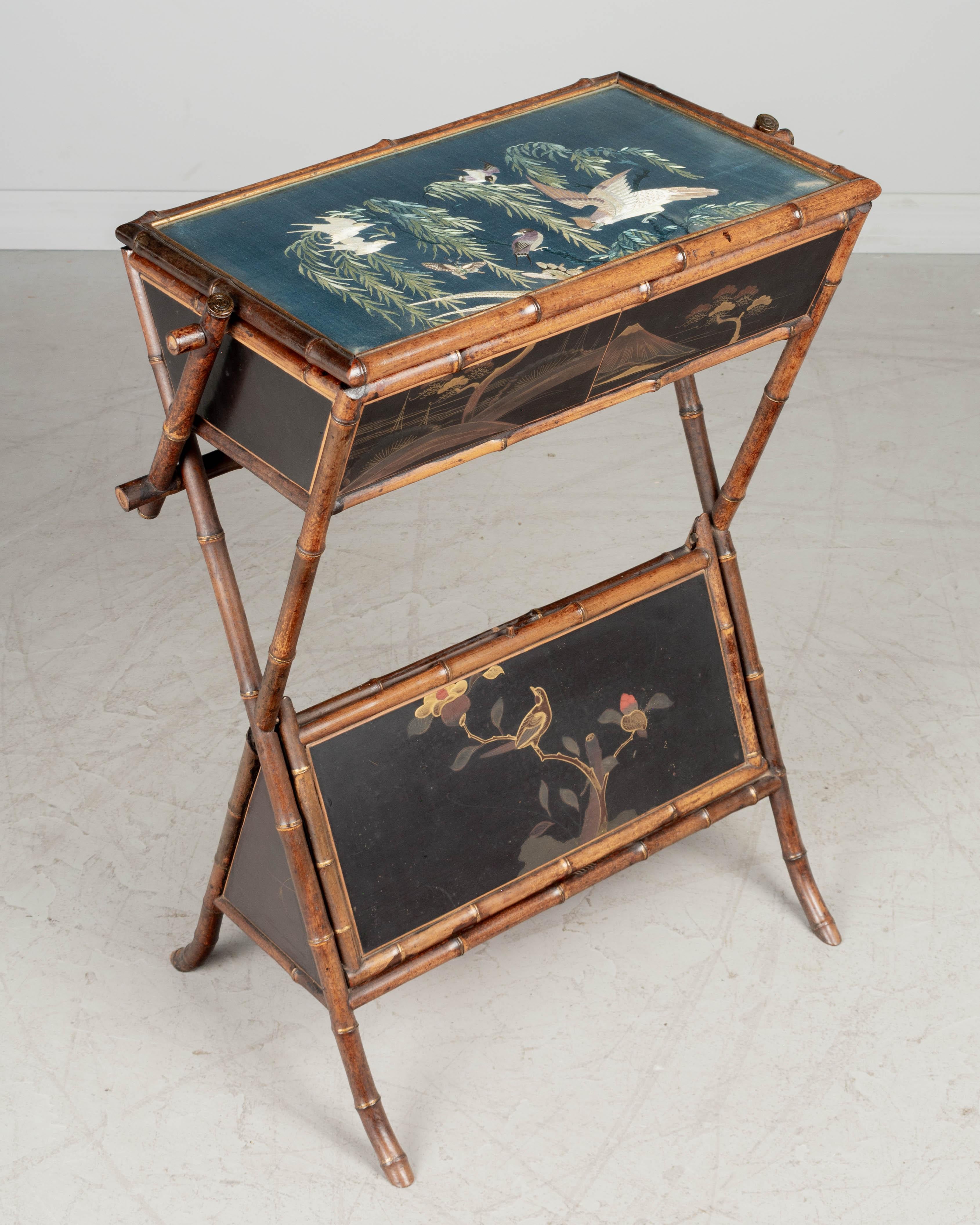 A 19th century French Japonisme Aesthetic Movement sewing table with bamboo frame and decorative black lacquer panels with red and gold cranes. Hinged top has a beautiful embroidered silk panel protected under glass a surface and opens to a mirrored