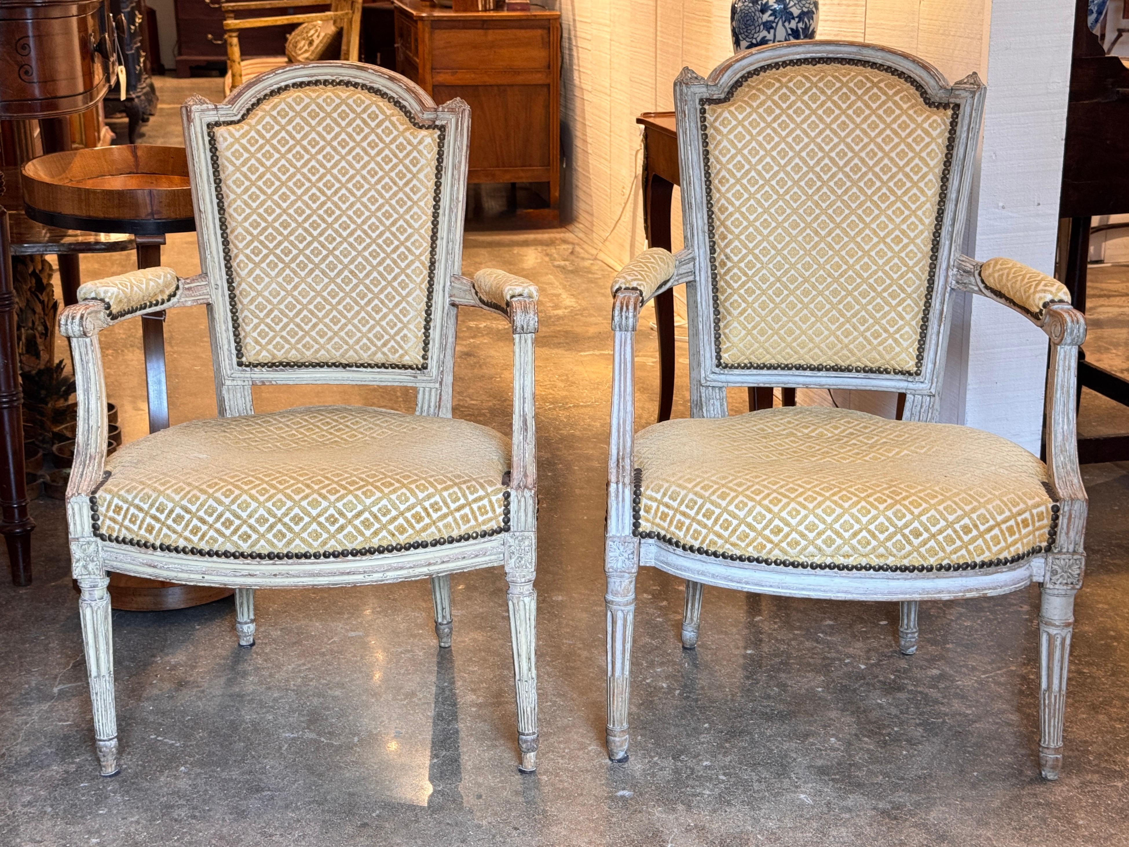 Pareja de sillones franceses del siglo XIX con respaldos perfilados, brazos con volutas y detalles florales tallados en toda la pieza. Los marcos de madera pintada descansan sobre patas estriadas. Los asientos y respaldos están acabados en un tejido