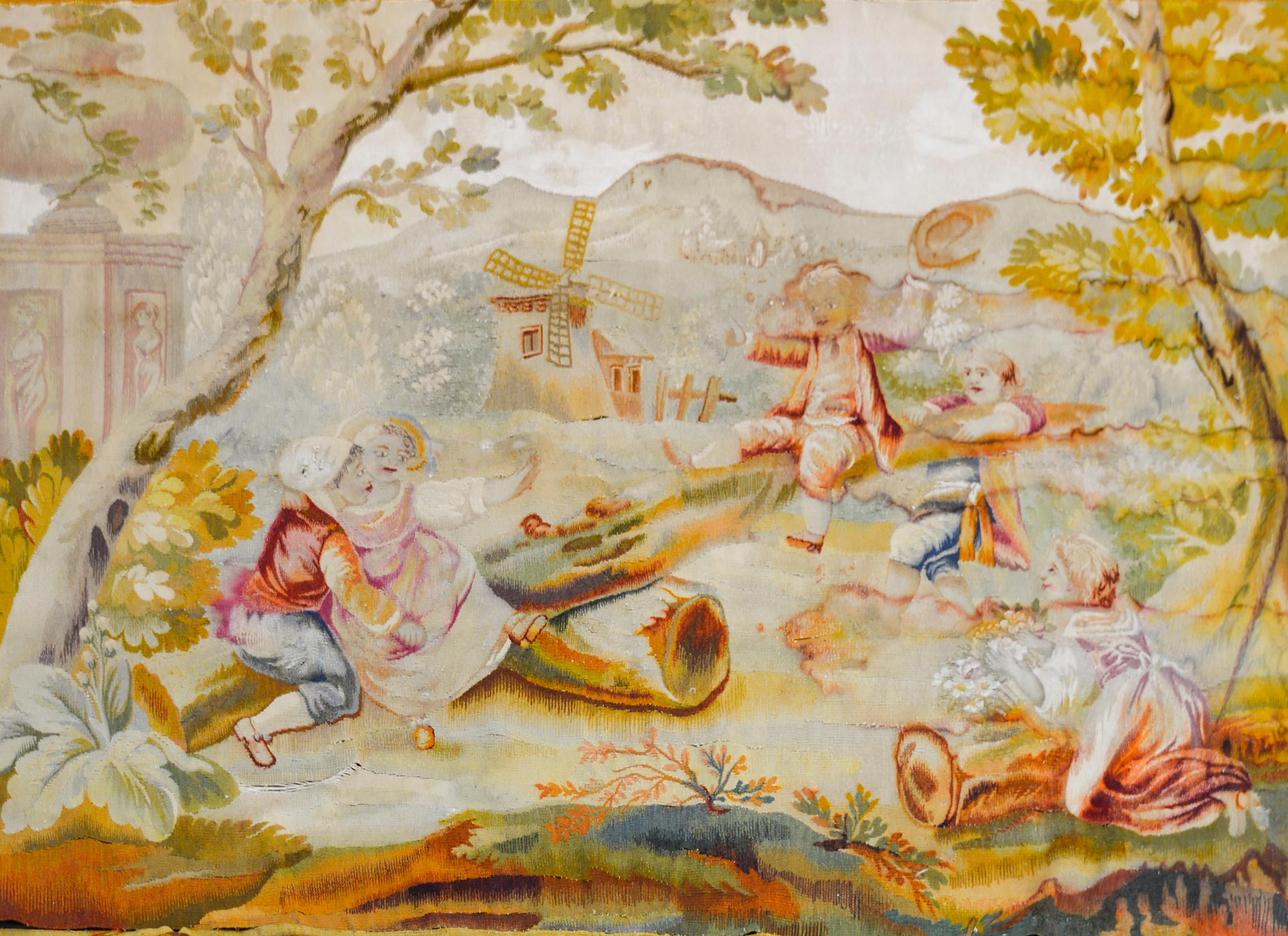 A wonderful mid 19th century French Aubusson tapestry depicting several children in period clothing playing on a log teeter-totter under an oak tree, and with a windmill and mountains in the background. The border is bold with a large-scale acanthus