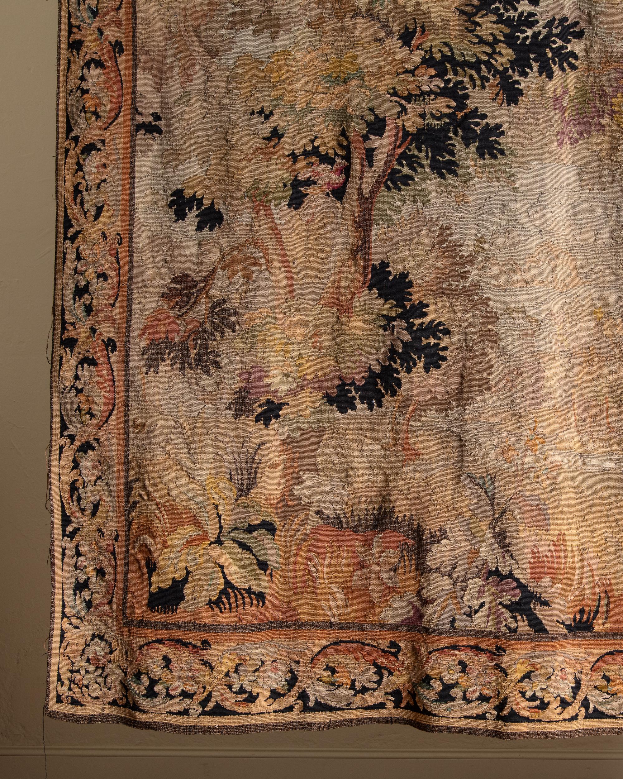 This is a 19th-century French Aubusson-style verdure woven tapestry, showcasing the refined artistry characteristic of the period. The tapestry depicts a lush woodland scene, featuring tall, leafy trees, dense foliage, and soft rolling terrain,