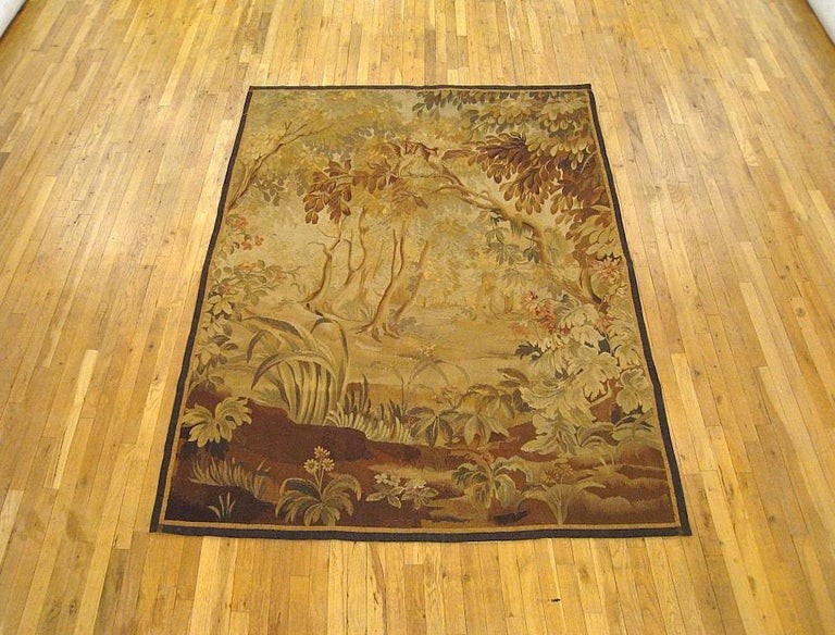 A French Aubusson verdure tapestry from the 19th century, depicting a woodland setting with two birds sitting on a tree branch above a variorum of bushes, flowers, and acanthus plants, and other trees in the distance. Enclosed by a pair of narrow