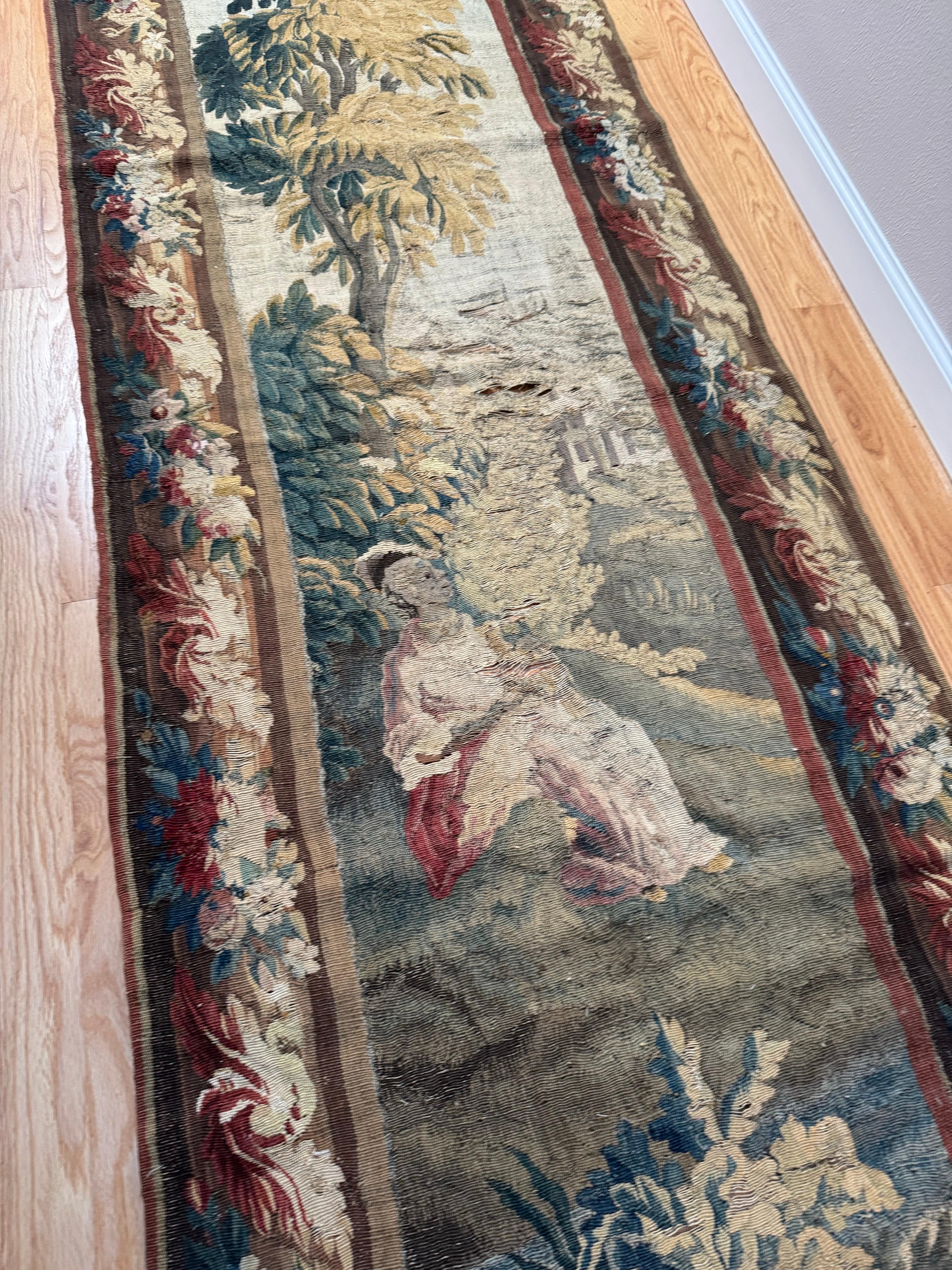 Place this long and narrow antique tapestry in a staircase or in between two windows for a pop of color. Handwoven in Aubusson, France in the 19th Century, this lovely verdure wall piece has its original fruit and floral border. It measures 8.9 x