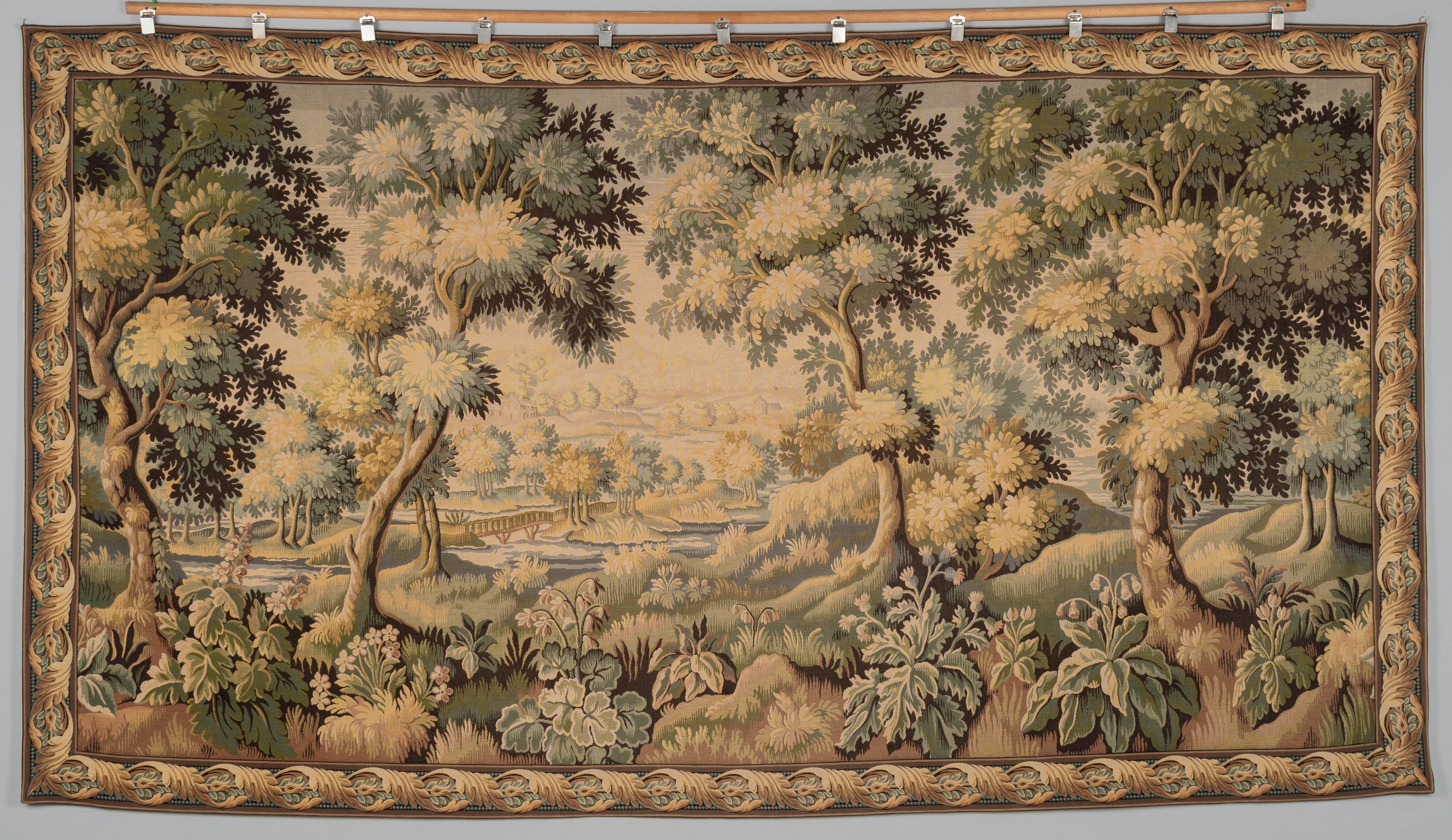 Ein großer französischer Wollteppich im Aubusson-Stil oder ein Wandbehang. Große horizontale Waldszene mit dekorativer Umrandung, großen Bäumen und Laub im Vordergrund, einem Bach mit Steg und einem Anwesen in der Ferne. Mechanisch gewebte Wolle mit