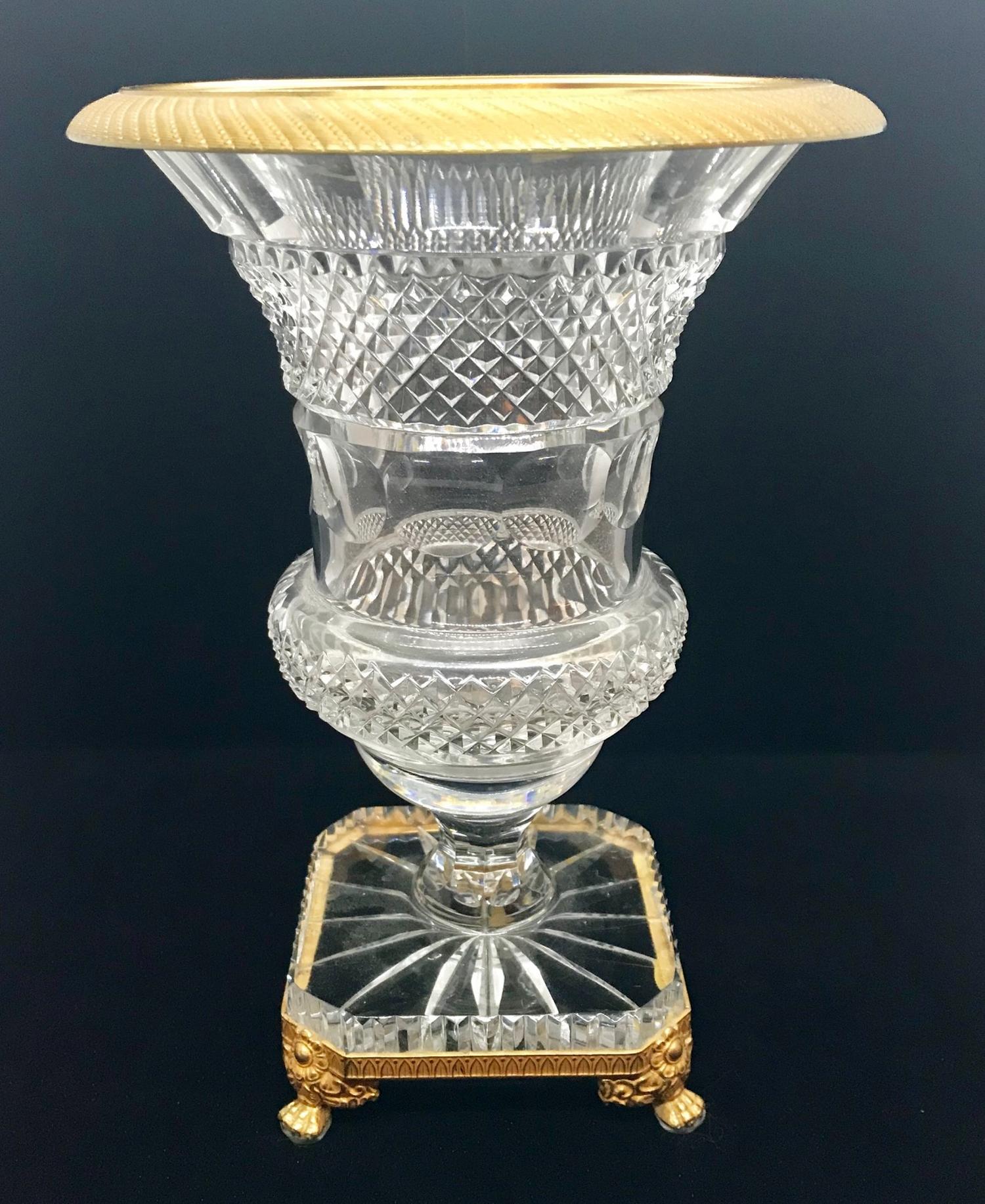 This fine cut crystal vase has a square fitted base which is bronze mounted and raised on four richly decorative feet. The ormolu rim is chased in a fine ribbon design. The baluster shaped urn is cut to perfection in round and diamond shapes.
