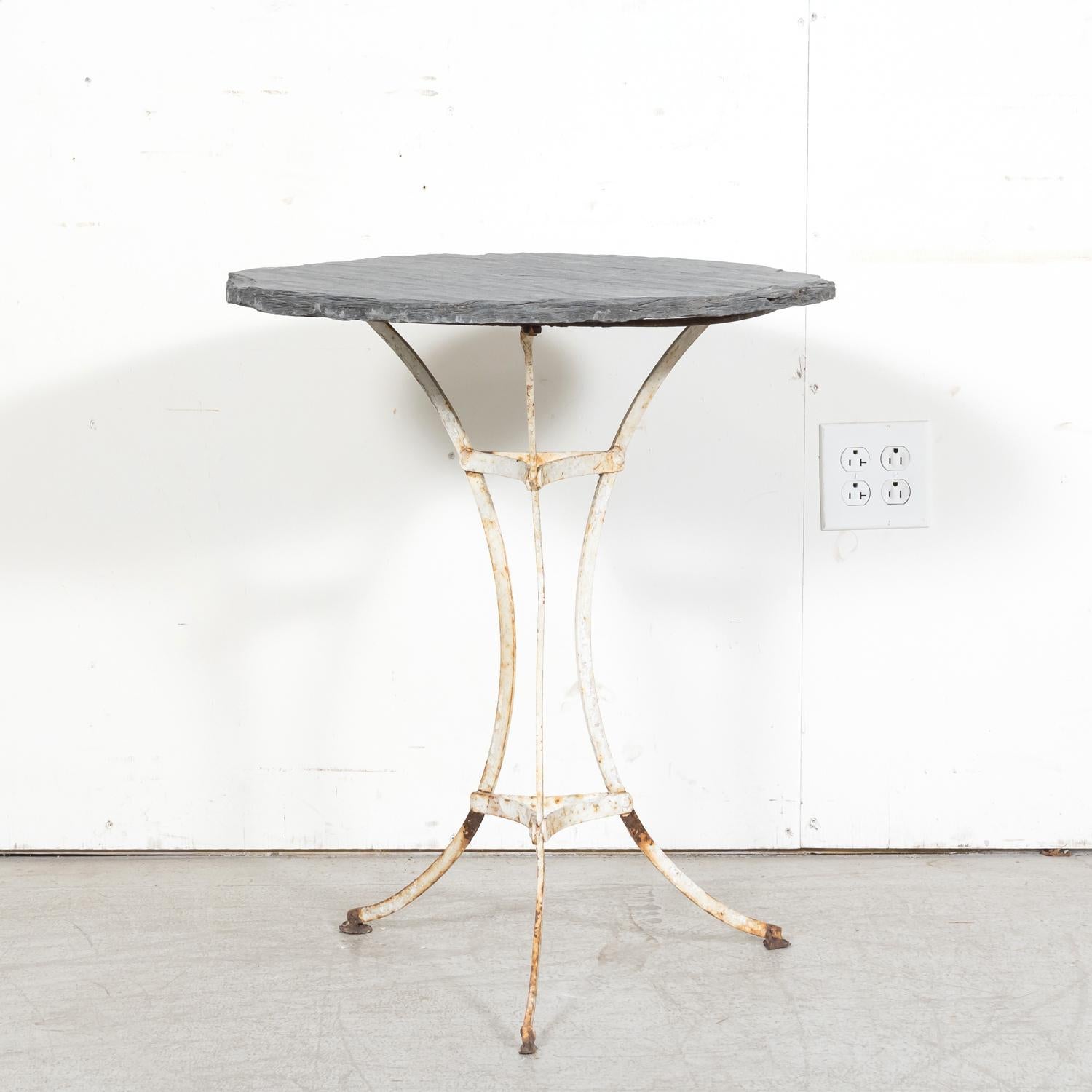 A 19th century French cast iron bistro or garden table, circa 1880s, featuring a slate top resting on a white painted iron tri-form base. The base boasts three scrolled legs joined with a circular iron stretcher, adding both stability and visual