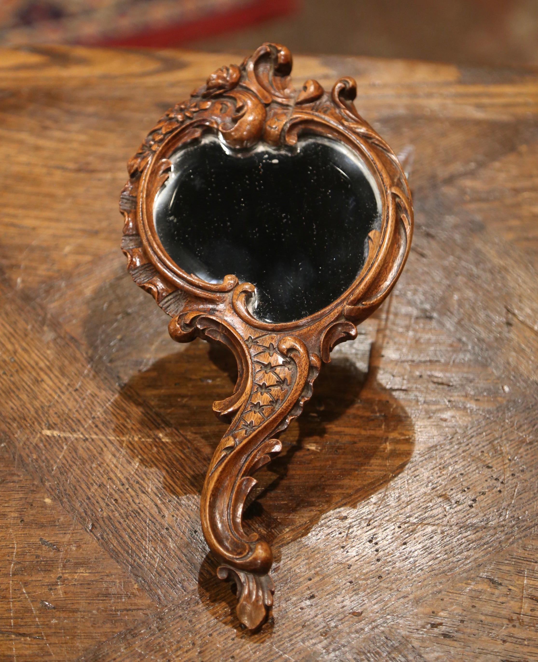 Crafted in France circa 1870, the antique Louis XV style hand mirror with elegant serpentine handle, features hand carved floral and leaf motifs throughout the frame; the vanity mirror has the original beveled mercury glass. The petite table mirror