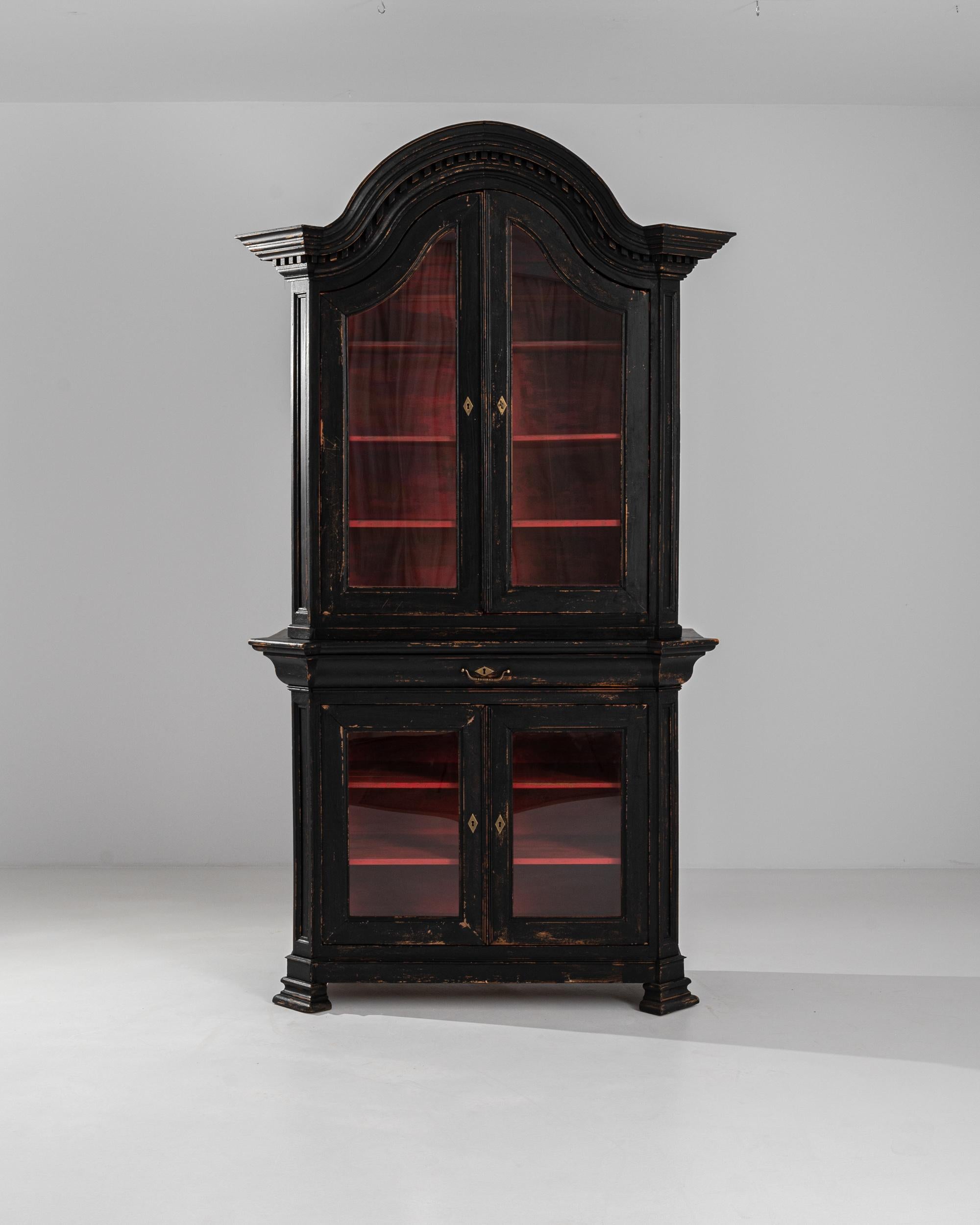 A painted wooden vitrine from 19th century France. Black and moody on the outside, and glowing vibrant red on the interior, this glass display case presents a complex and intriguing persona. Neoclassical moldings and a dramatic arched shape announce
