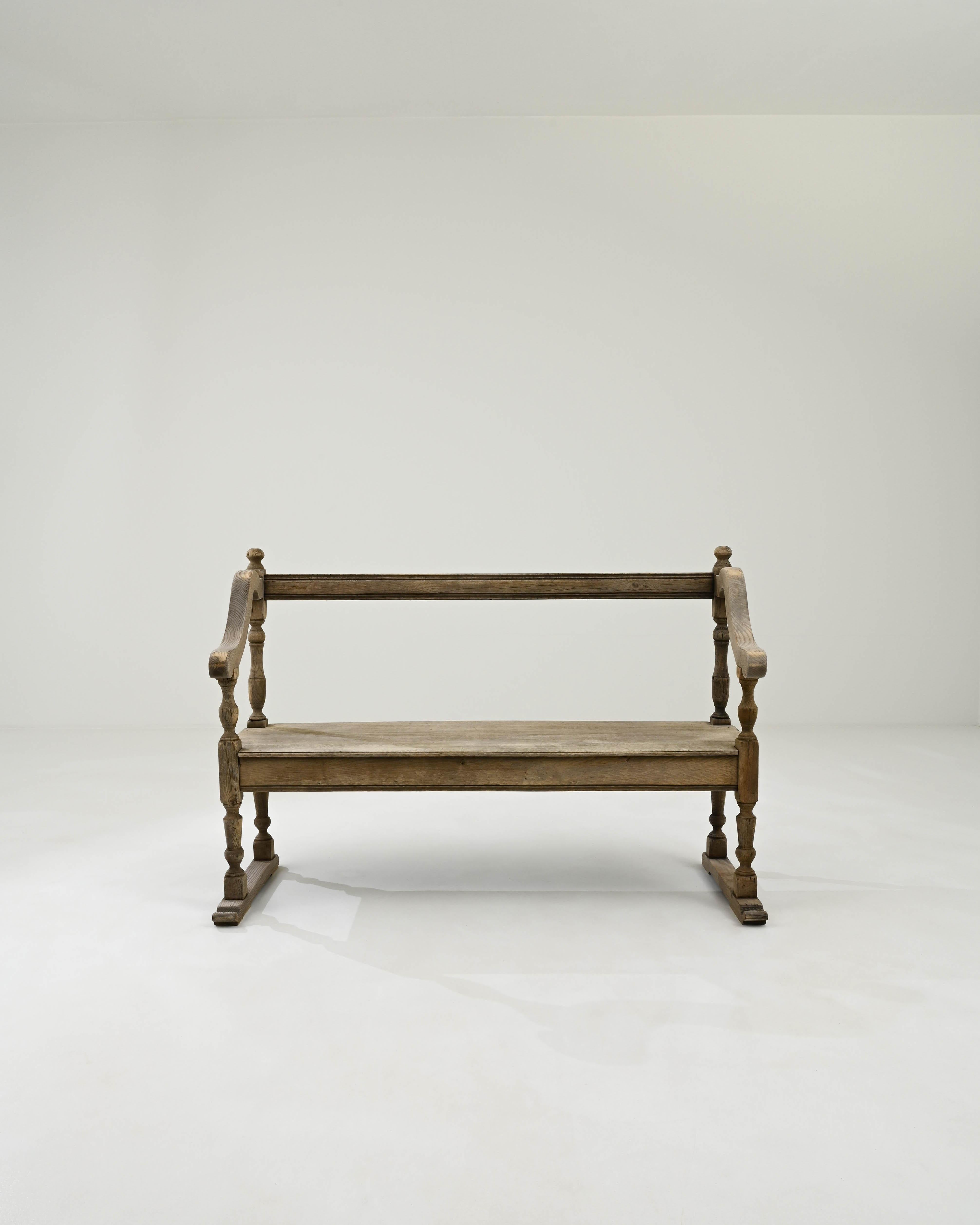 Hand-crafted from fine French oak, this 19th-century bench showcases artfully carved baluster legs that culminate in a stylish base, providing extra support and lending the piece a distinctive appeal. The playfully curving armrests are adorned with