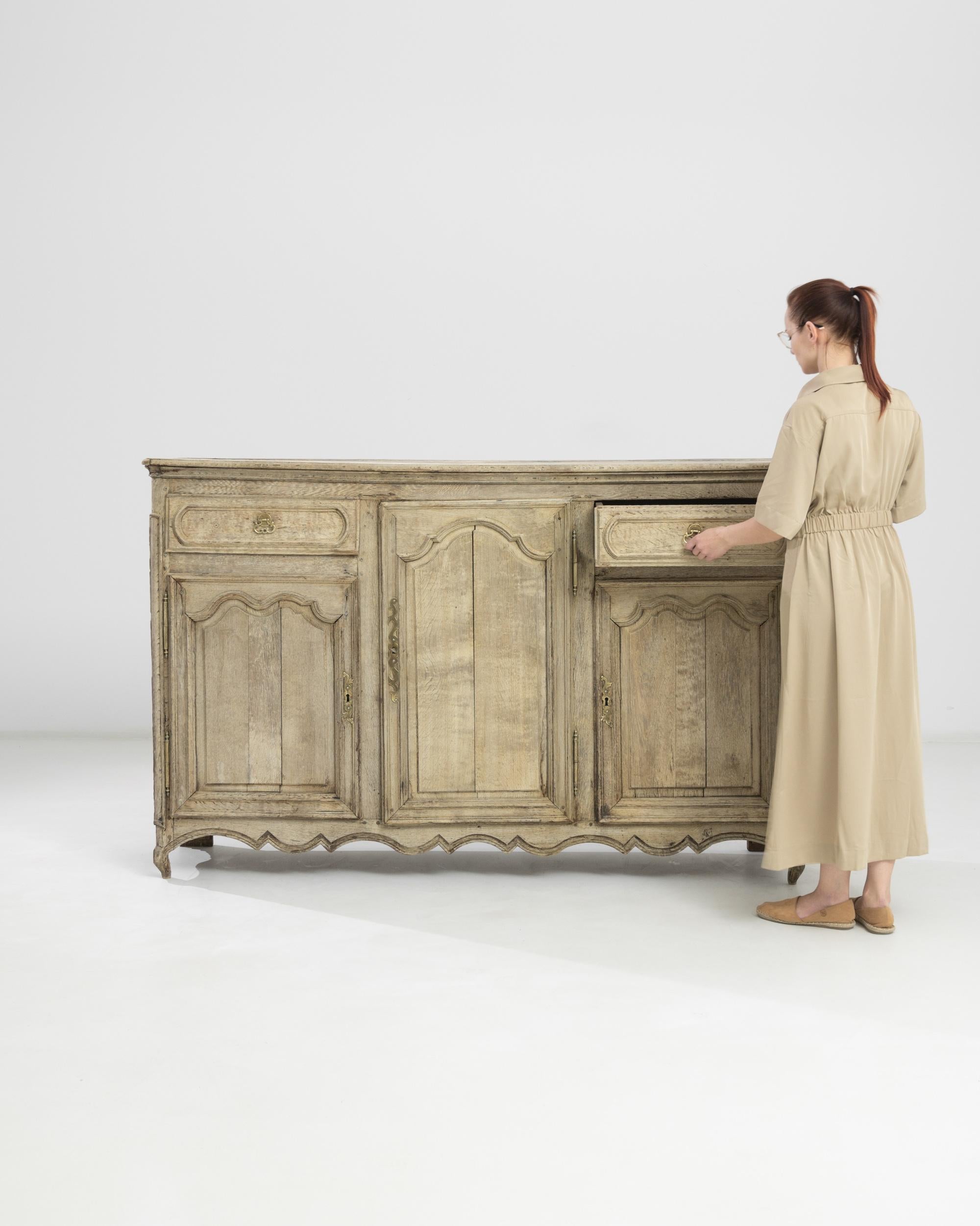 Crafted in France circa 1860, this bleached oak buffet flaunts gentle curvilinear arch carvings on the raised doors, echoed in the waves of the scalloped apron and the bend of its miniature convex legs. The original gilded hardware of the drawers