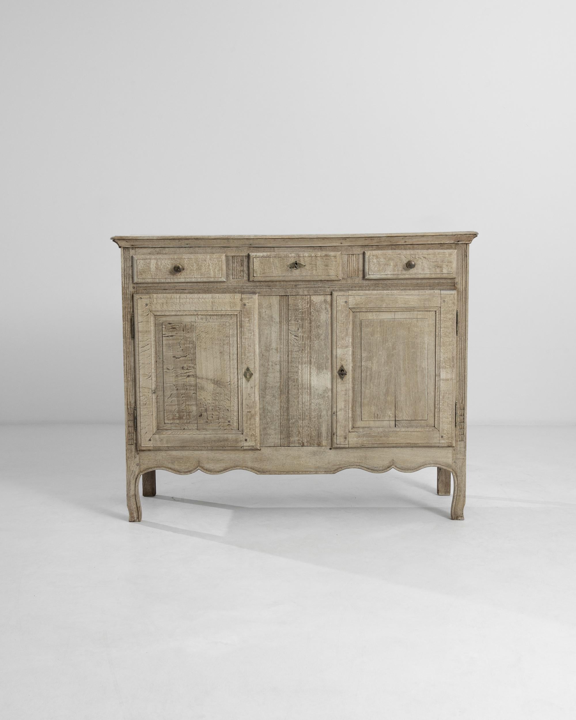 A 19th century bleached oak buffet made in France. Modestly decorated with a scalloped skirt and molded cornice, the chest features three shallow drawers and two square panel doors held up by a pair of slightly curvy front feet. The dim shining of