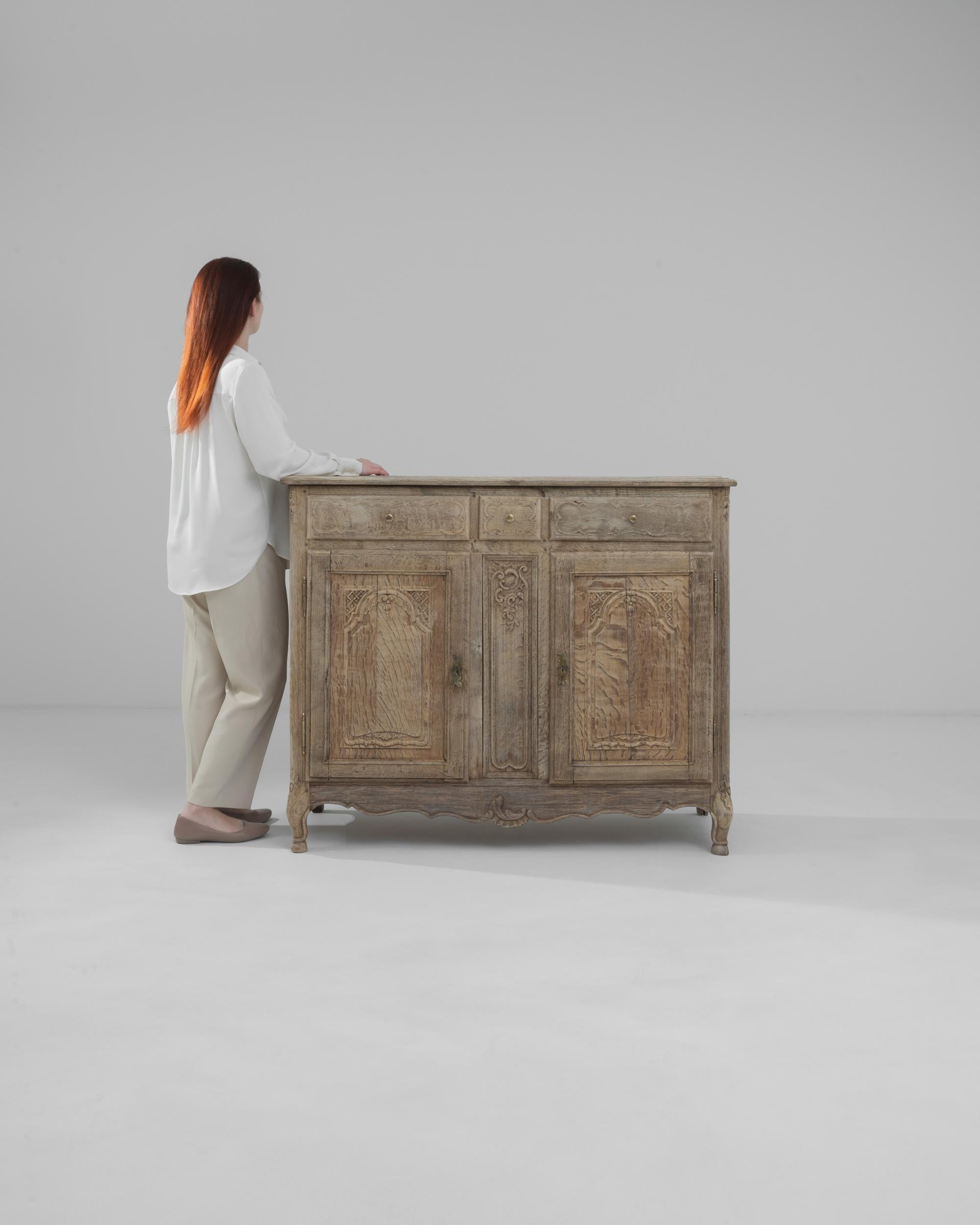 This 19th Century French Bleached Oak Buffet exudes a stately presence, with its robust and time-worn facade evoking tales of grandeur from a bygone era. The bleached oak finish highlights the natural wood grain and intricate carved details, giving