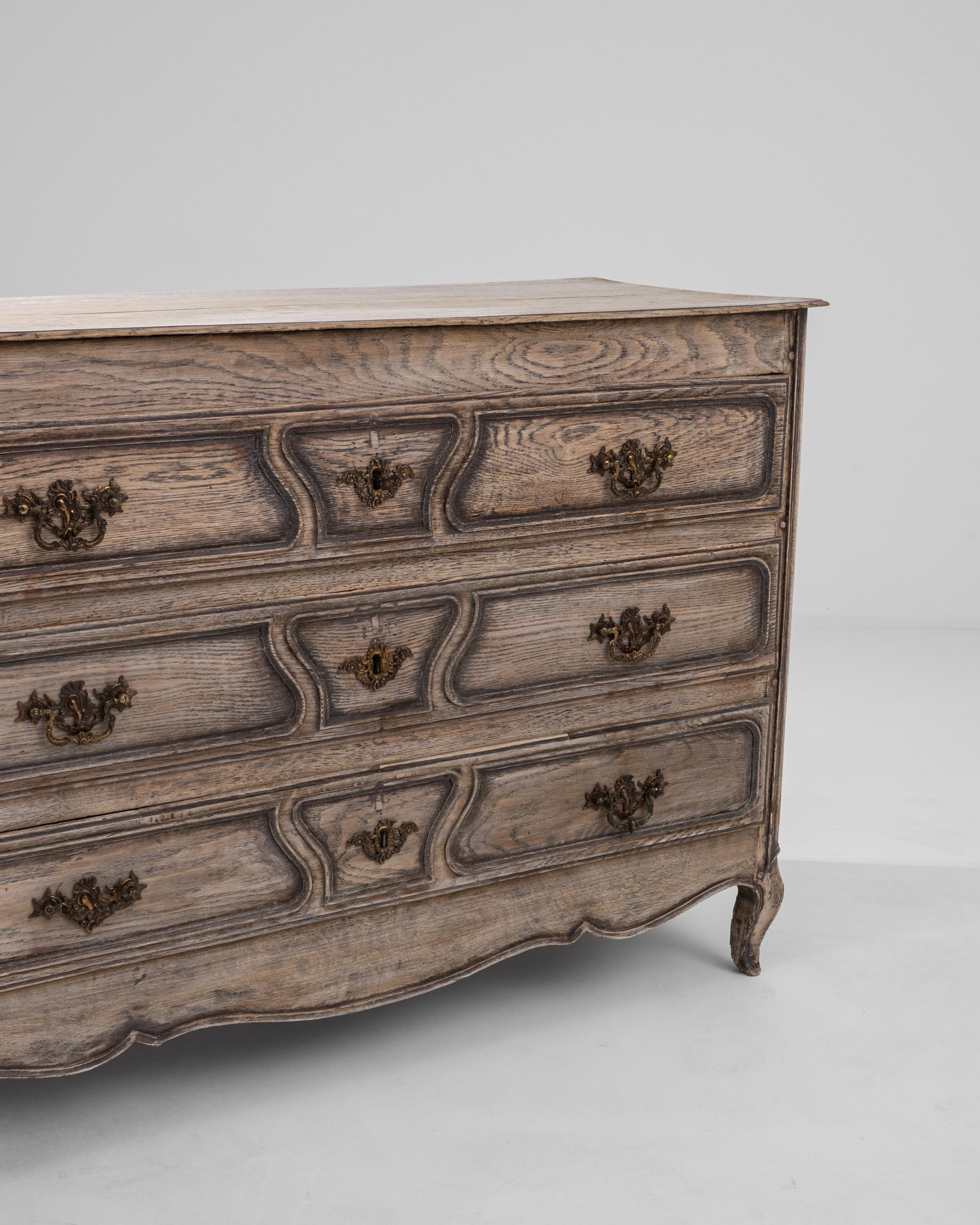 Embark on a journey of refinement with this 19th Century French Bleached Oak Chest of Drawers. Exuding timeless elegance, this piece features three spacious sliding drawers adorned with intricate engraved patterns. The scalloped apron and front