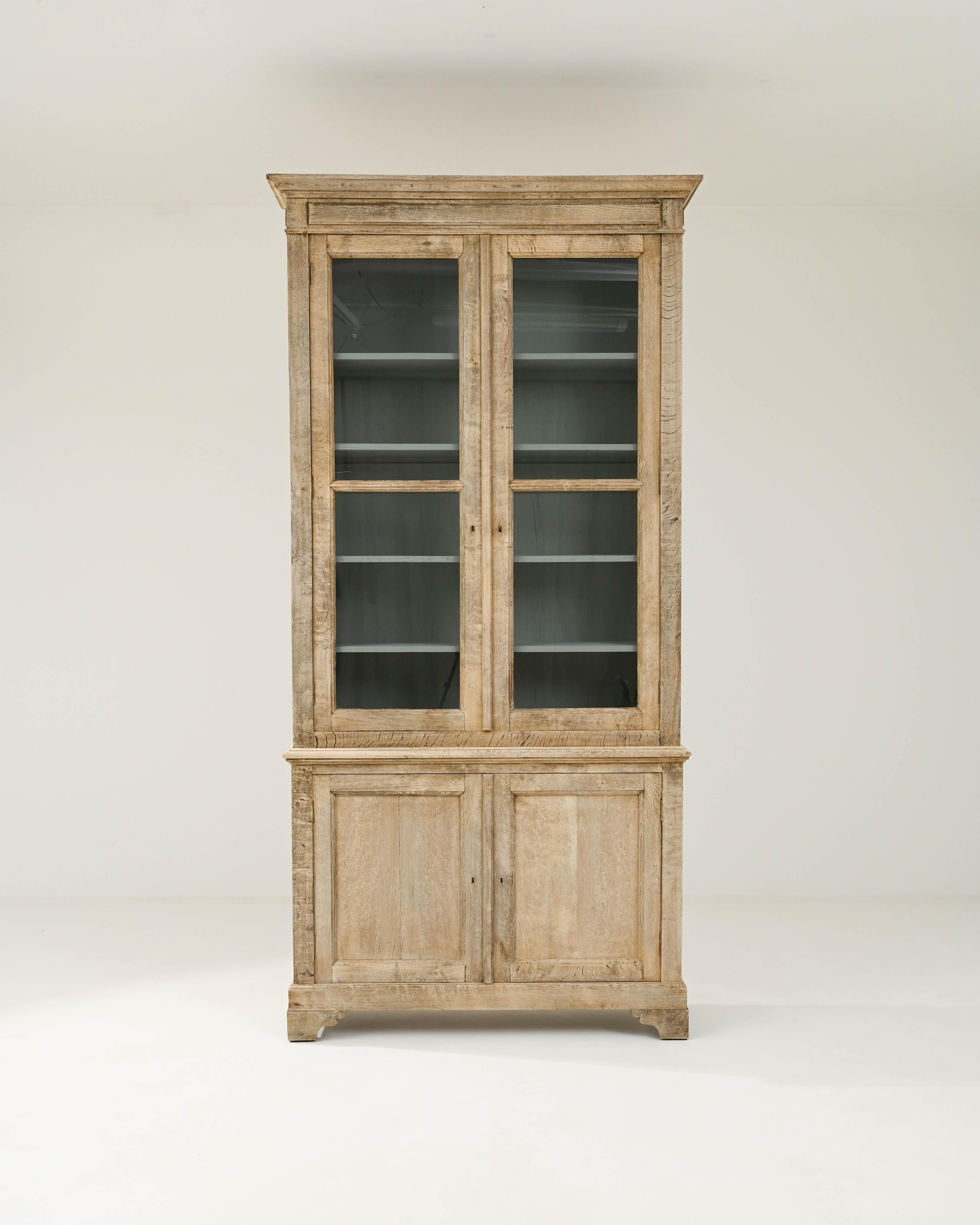This 19th-century à deux corps vitrine commands attention with its grandeur and understated elegance. The towering stature of this French cabinet is emphasized by a molded cornice with pointed corners. Expertly carved bracket feet and classical