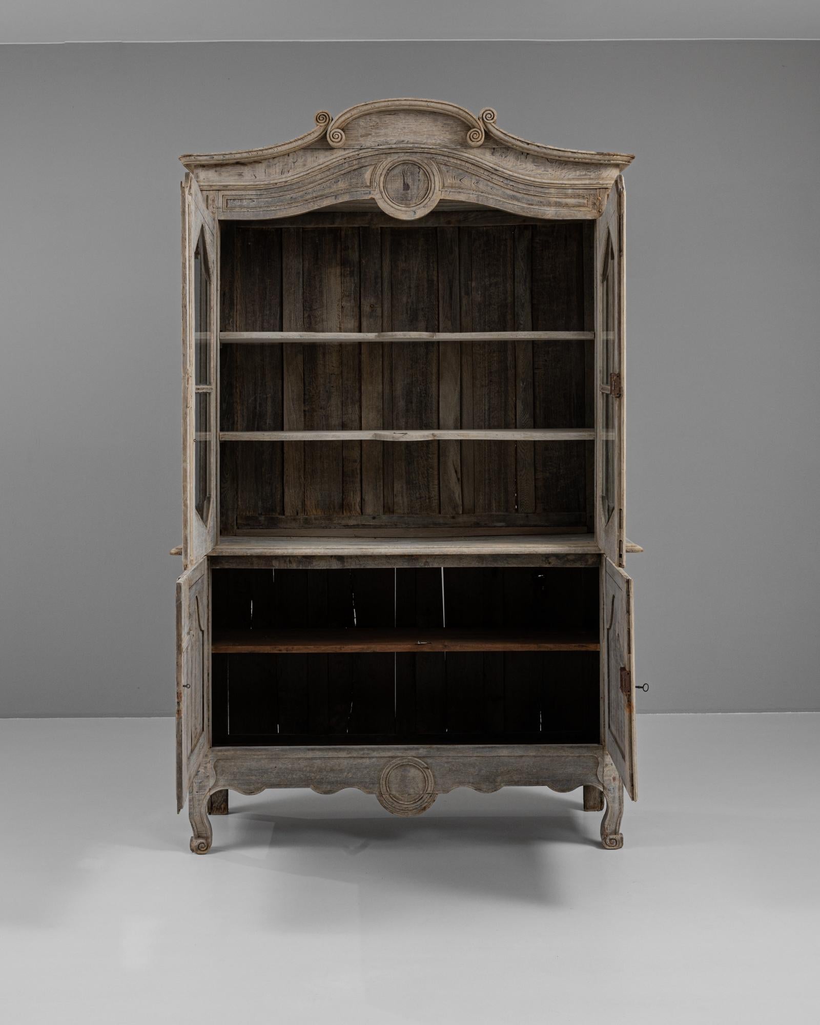 This exquisite 19th-century French vitrine, crafted from bleached oak, exudes elegance and antique charm. Featuring intricate carvings along the arched crown and cabinet doors, this piece showcases the craftsmanship of its era. The vitrine's upper