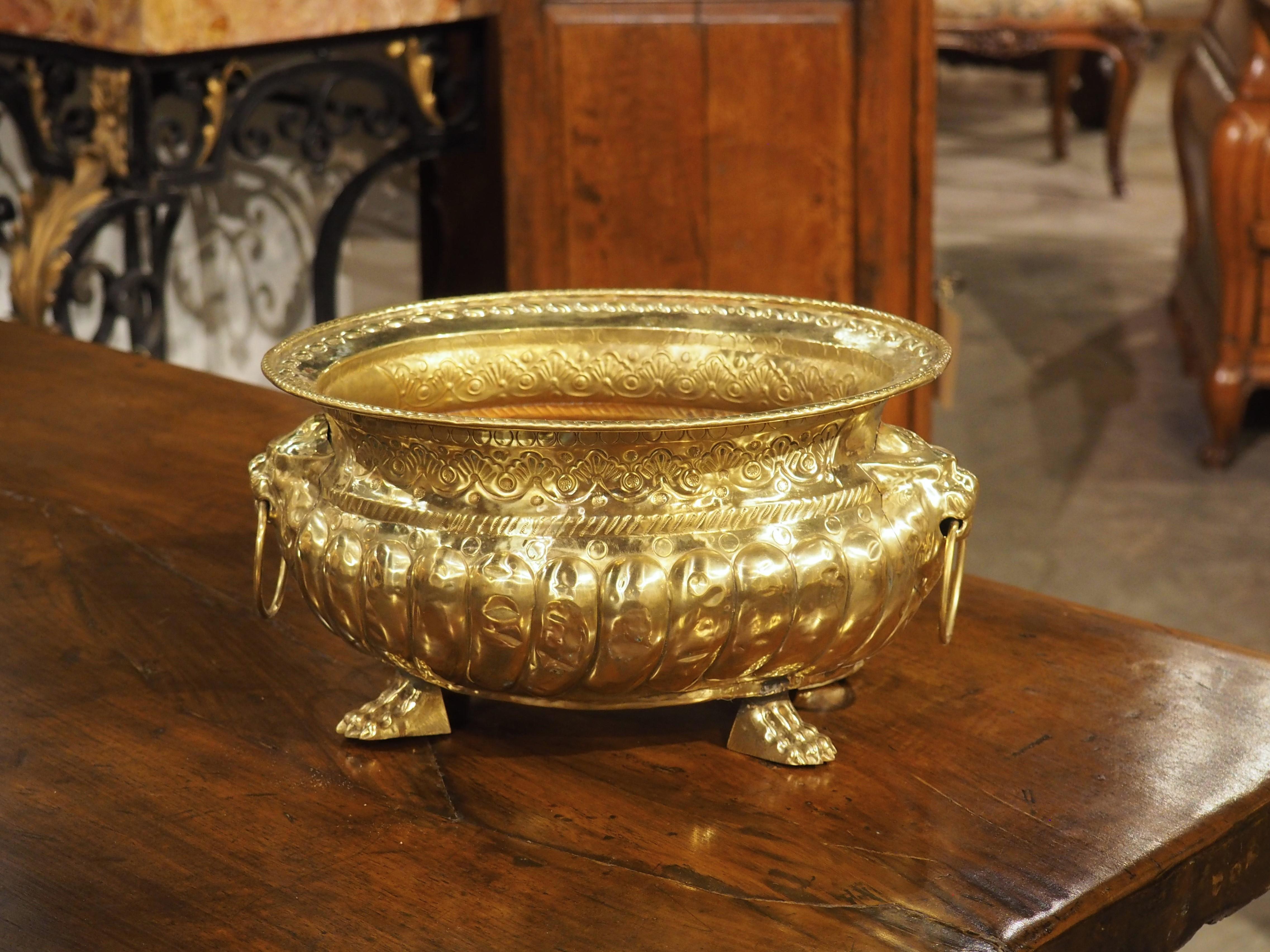 This ovate French brass jardiniere from the 1800s has thick lobes, accompanied by lion mascaron handles and four monopodial paw feet. The face of a lion on each of the sides interrupts the continuous gadroon of the main body. Both lion faces have a
