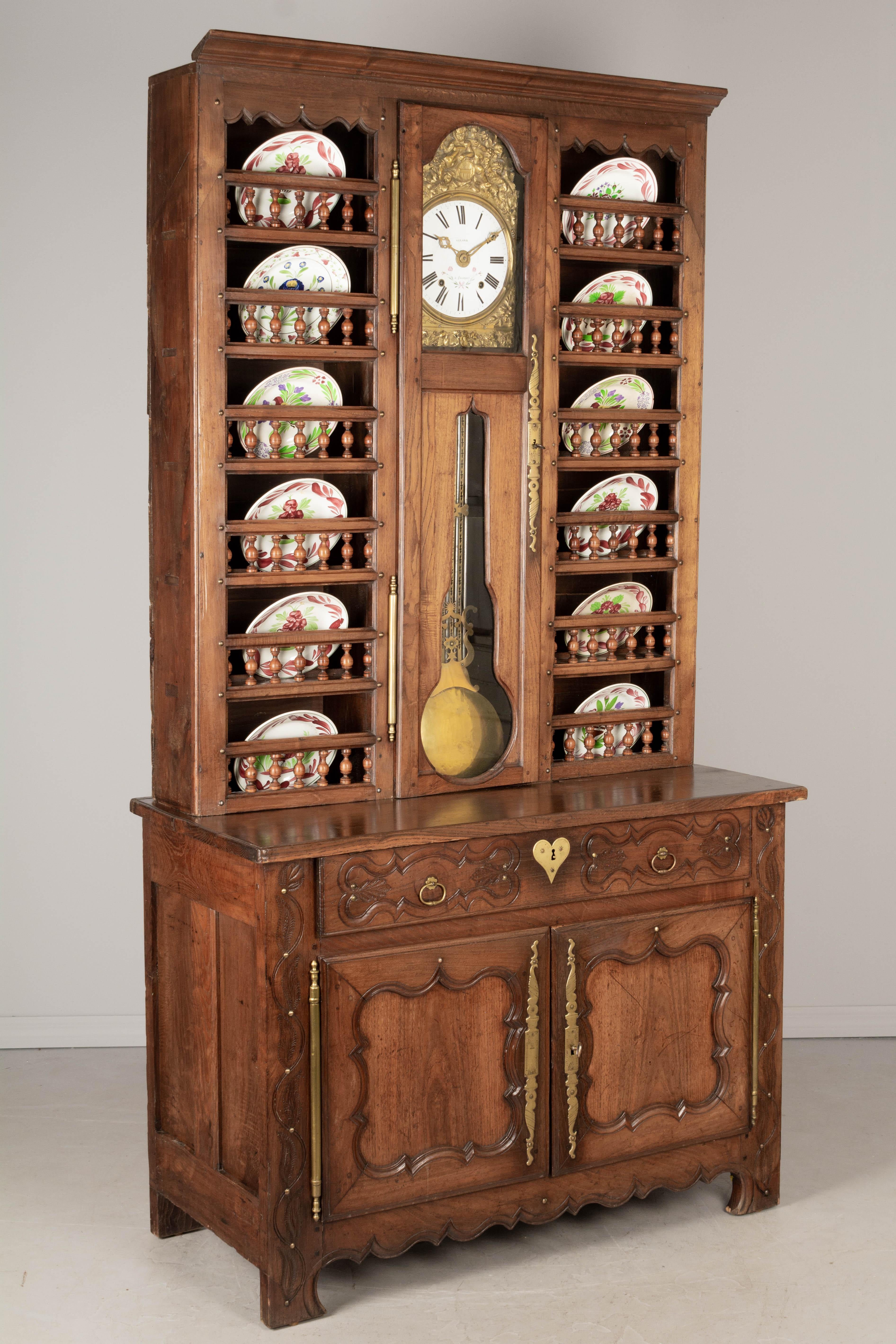 A 19th Century Country French vaisselier, or china hutch, from Brittany made of solid chestnut and in two parts. The top has a tall plate rack with two sets of six shelves with turned spindle gallery flanking a glass cased clock. Clock has an enamel