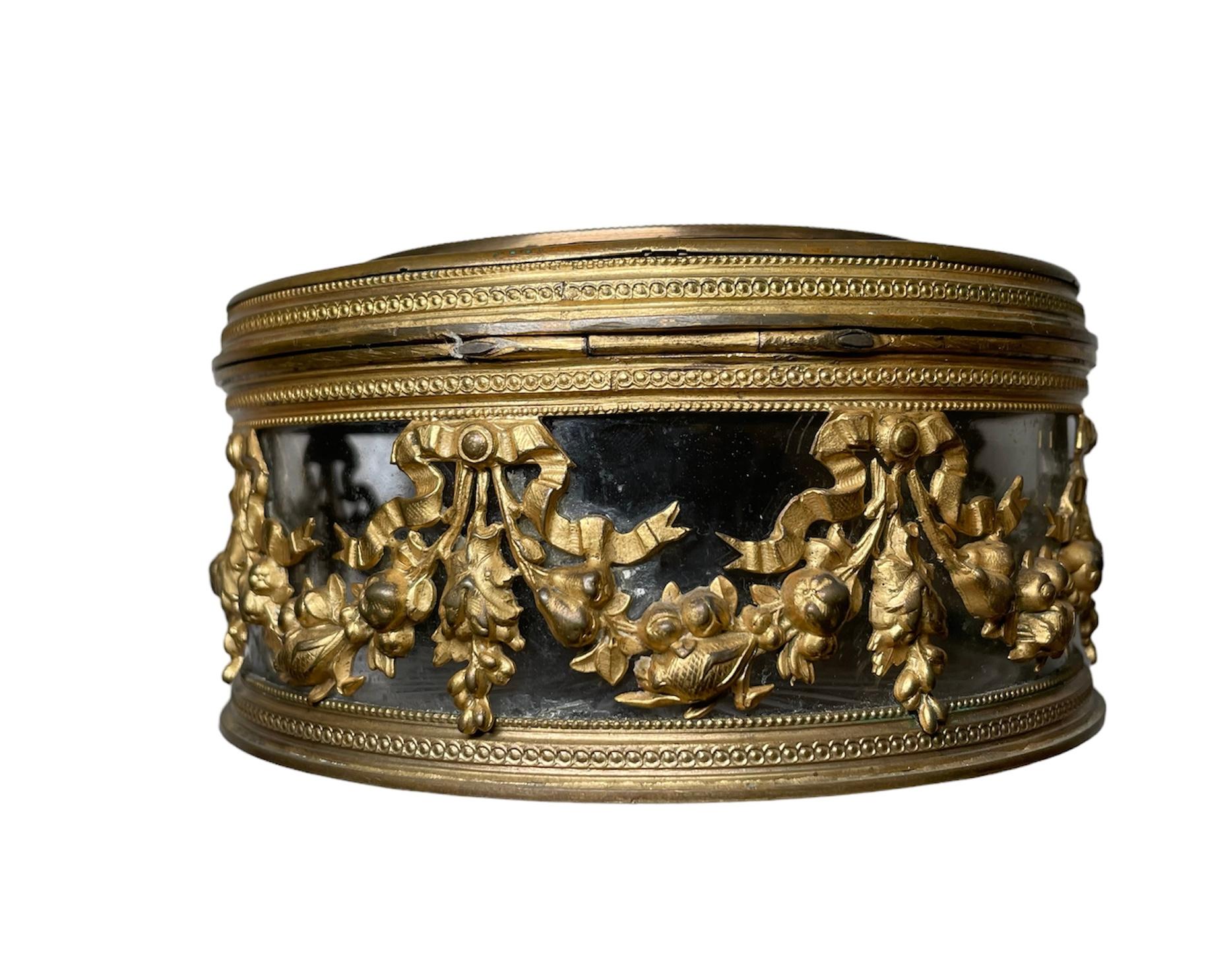 This is a French bronze metal overlay round powder glass jar/box. It depicts a hinged lidded glass jar decorated with a long gilt garland of bows and flowers and some fine beads. The lid is also adorned with the same, but it is enhanced with a hand