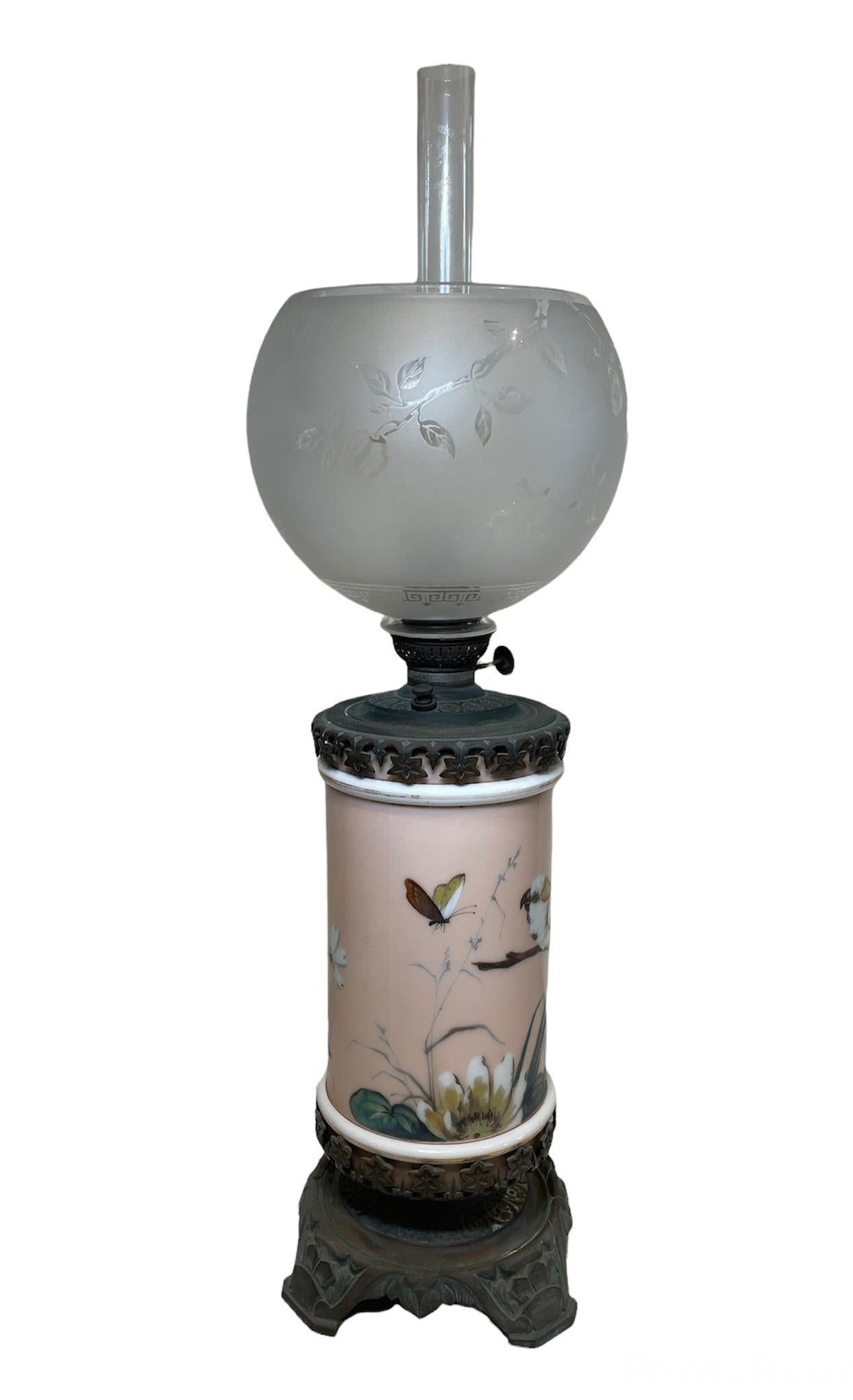 This is a hurricane oil lamp made of bronze and porcelain. It depicts a cylindrical shaped porcelain font hand painted with a light pink background embellished by some flowers and a nature scene of a cockatiel standing in a branch observing a