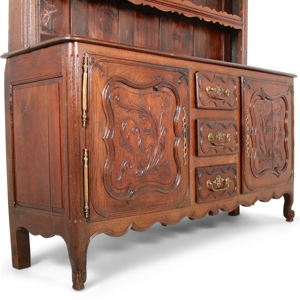 A 19th century French-country carved oak buffet, the base with two panelled doors flanking a bank of three central drawers, the top with three open shelves.
The lower doors have shaped serpentine frames with conforming panels, all hand-carved with