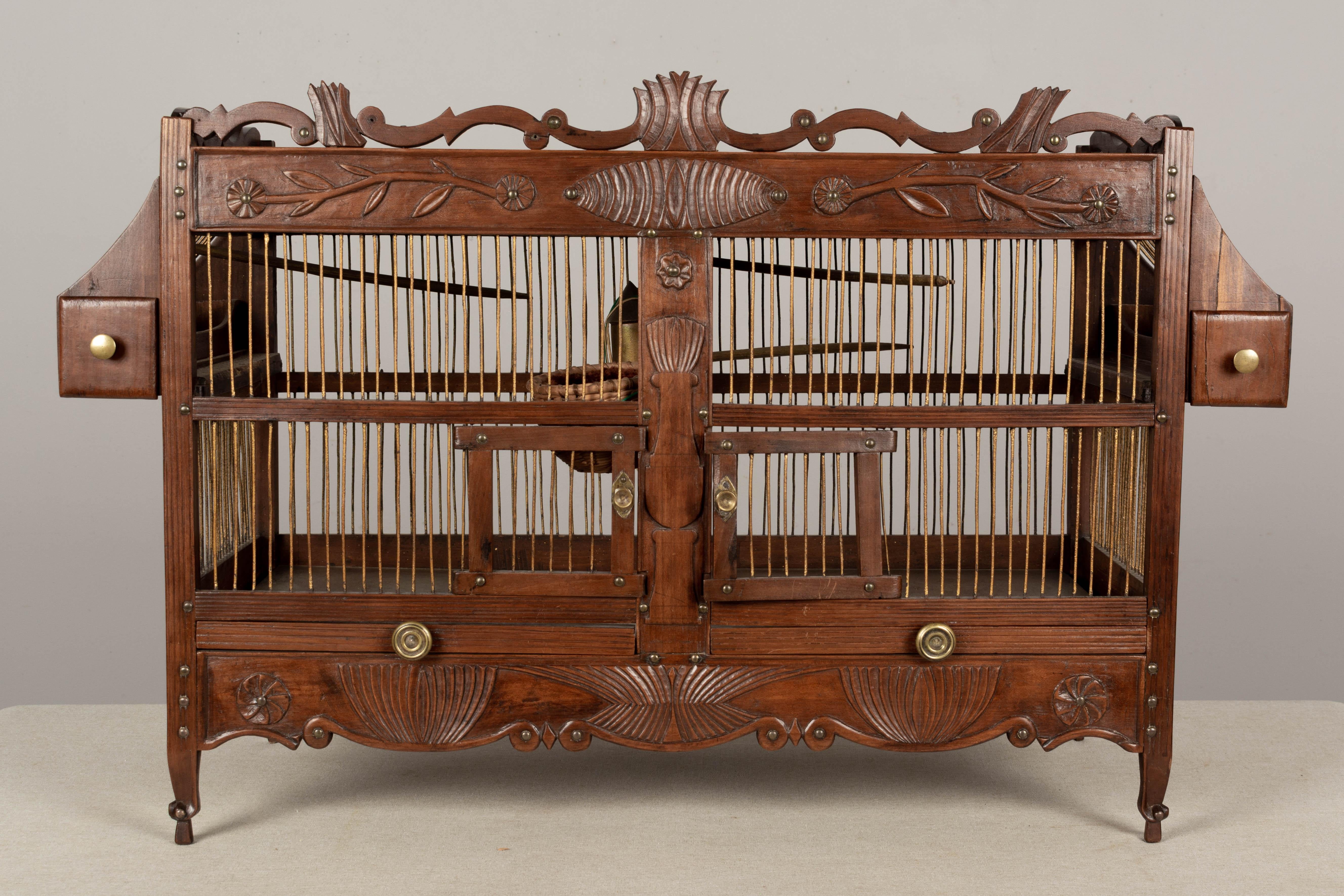 A 19th century French birdcage made of brass wire and cherry wood with hand-carved decoration and brass nail trim. Pull-out trays at the bottom with mirrored glass plates and pull-out seed troughs at the top, each with brass knobs. The interior is