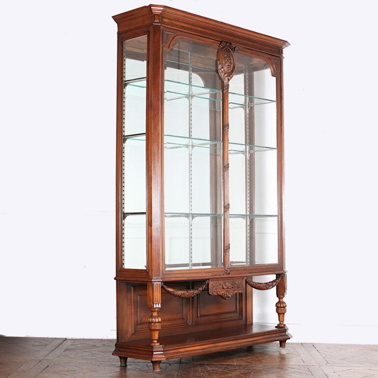 A grandly-scaled late 19th century French carved walnut Louis XVI style vitrine, the case with finely hand-carved swags, ribbons and floral details. Two doors retain their original shaped beveled glass; sides with original beveled glass also. Three