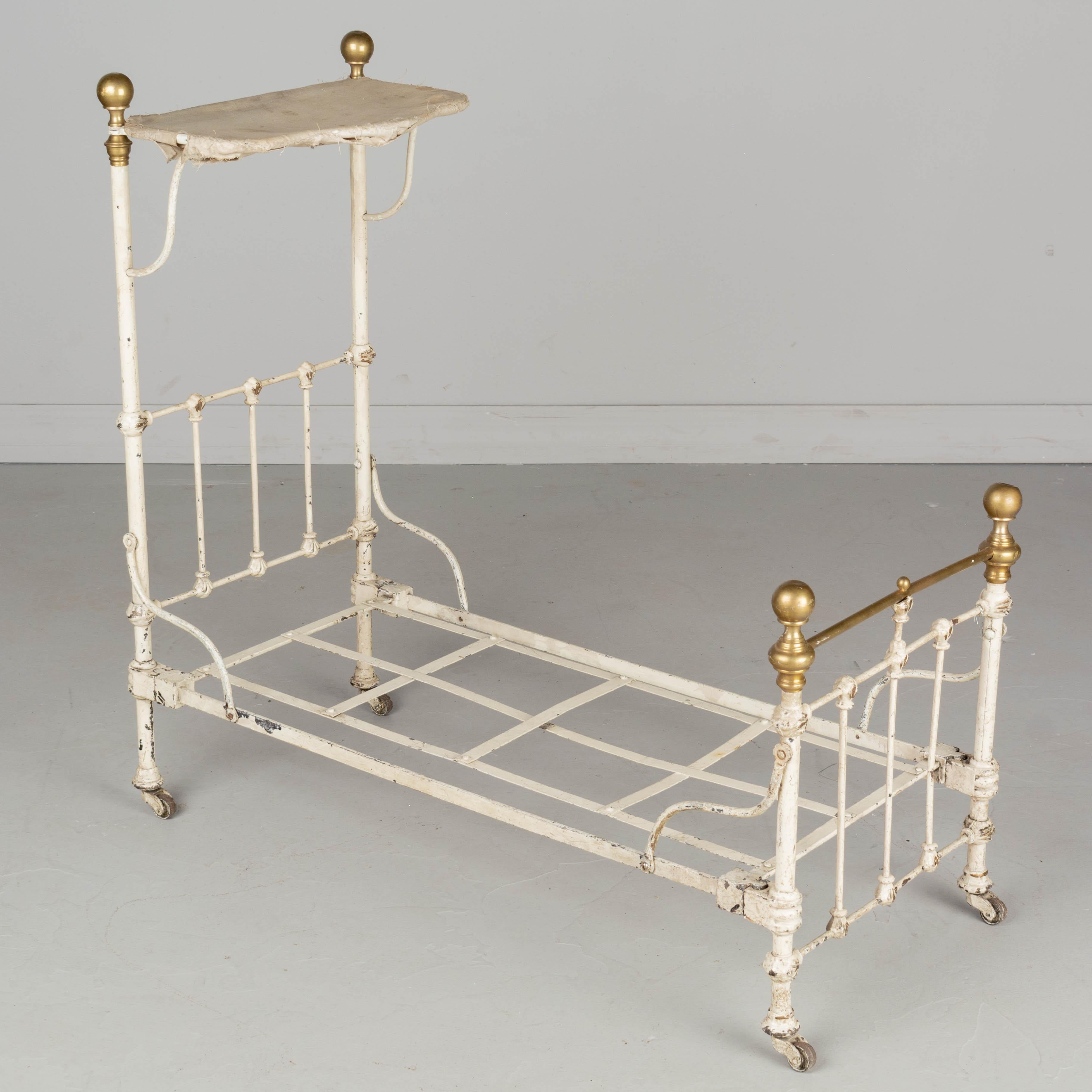 A French cast iron miniature sampler bed with white painted patina and brass finial decorative elements. Woven iron straps support a 13.5
