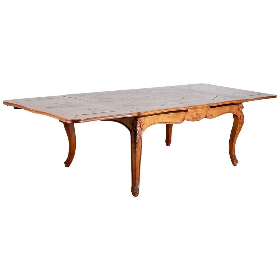 A late 19th century French country cherrywood 'drawleaf' dining table, the top and leaves with inlaid solid cherry parquet diamond pattern, the Louis XV style base with carved cabriole legs with scroll feet and carved knees, the serpentine profile